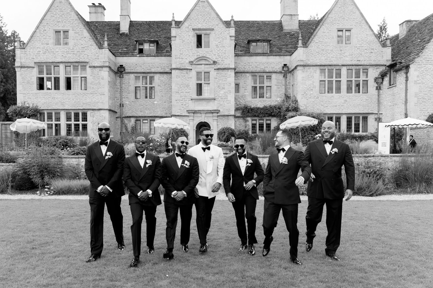 Groom and groomsmen in black tuxedos pose outside grand stone manor house.
