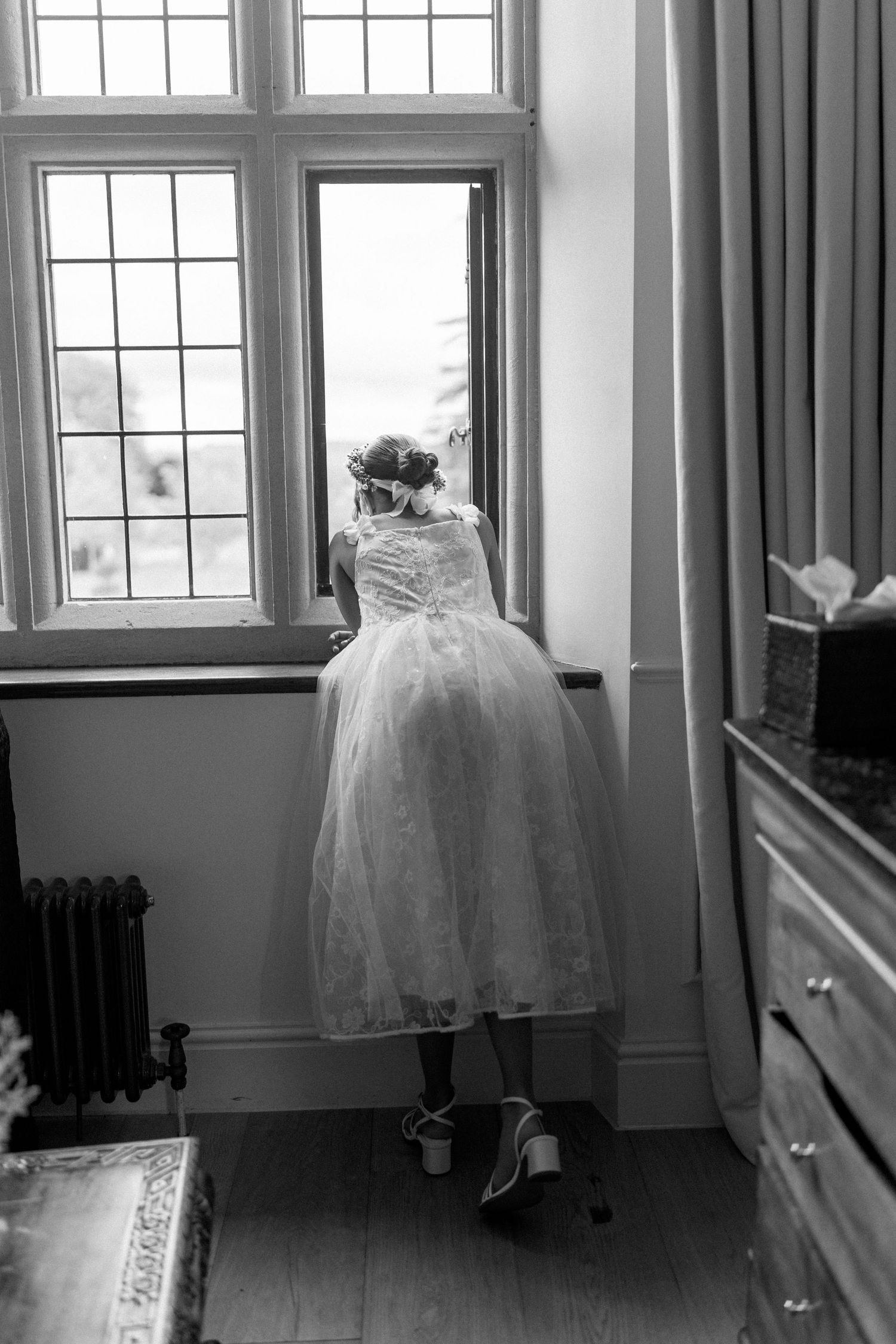 Flower girl in short lace dress peeks out open window in black and white wedding photo.