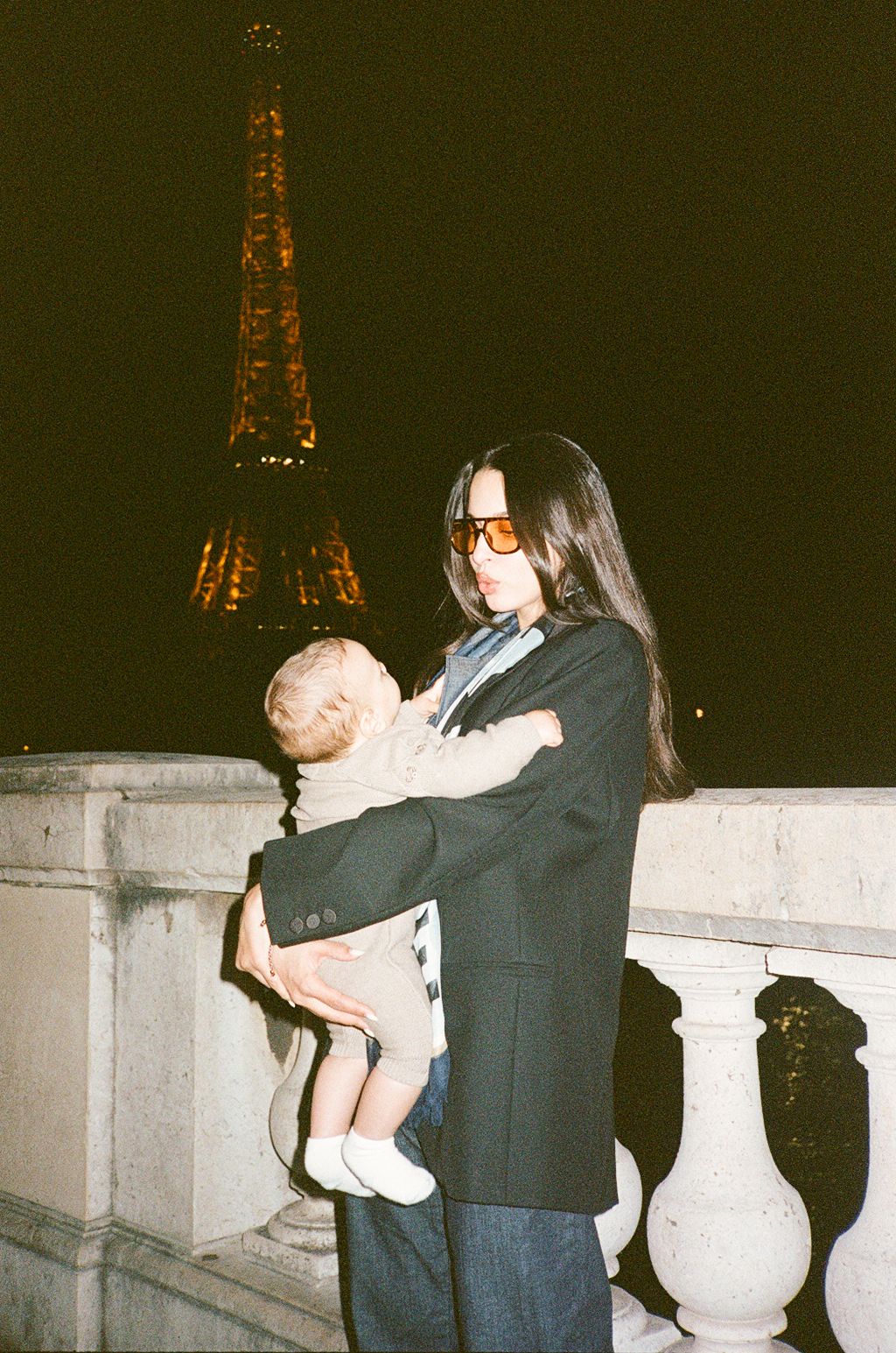 Woman in black blazer holding a baby in front of the illuminated Eiffel Tower at night in Paris.