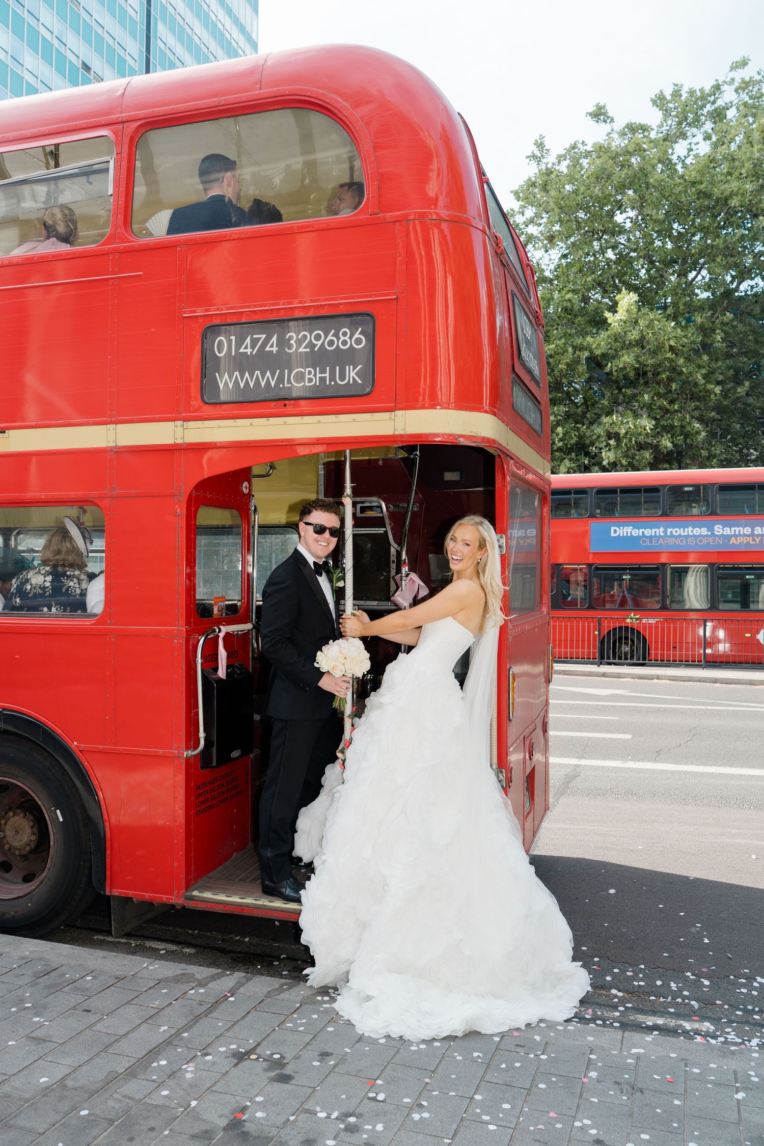 Bride and groom posing on a classic red London double-decker bus in wedding attire, holding a white bouquet.