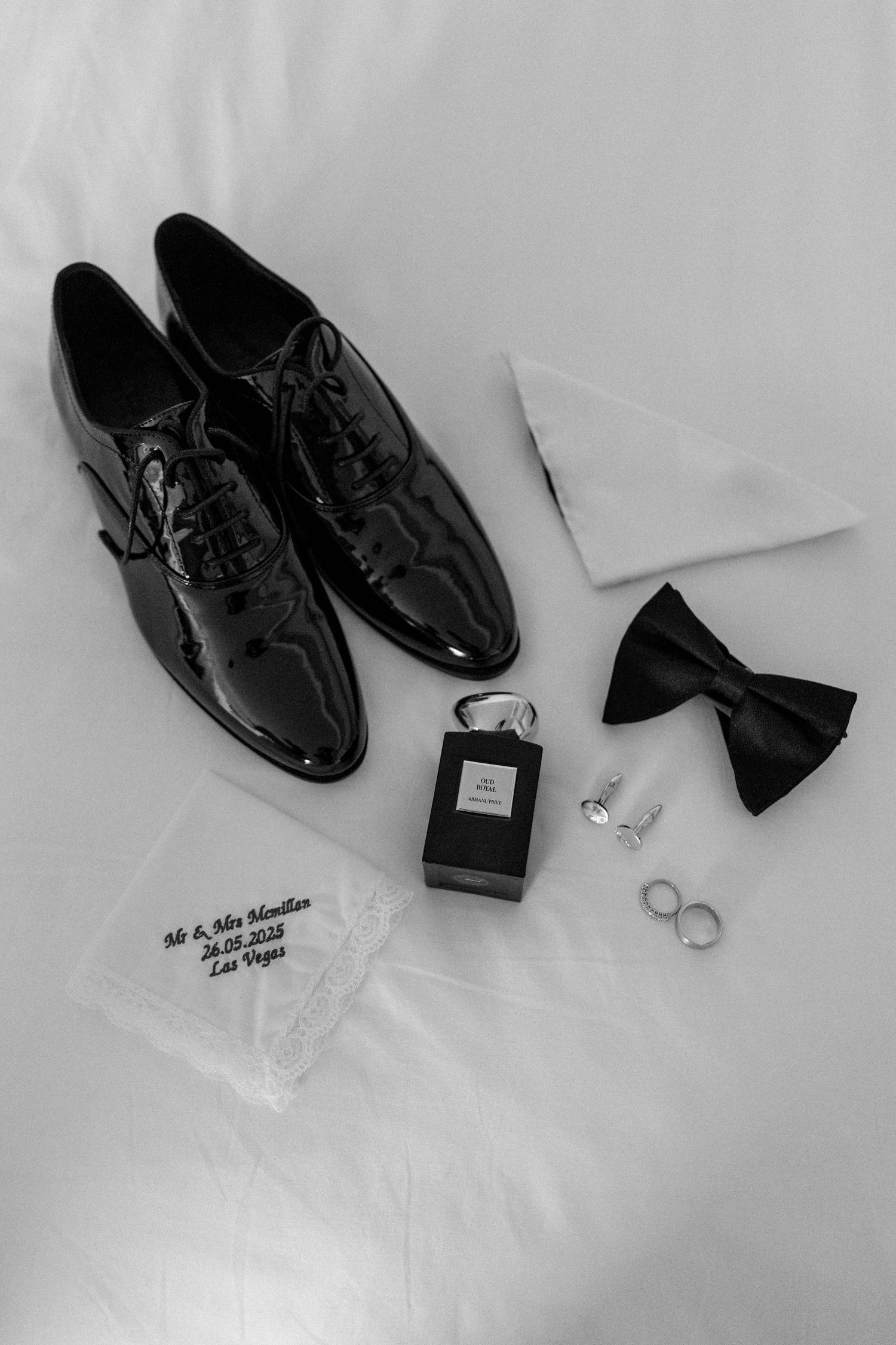 Wedding accessories including black dress shoes, rings, and bow tie arranged on white surface.