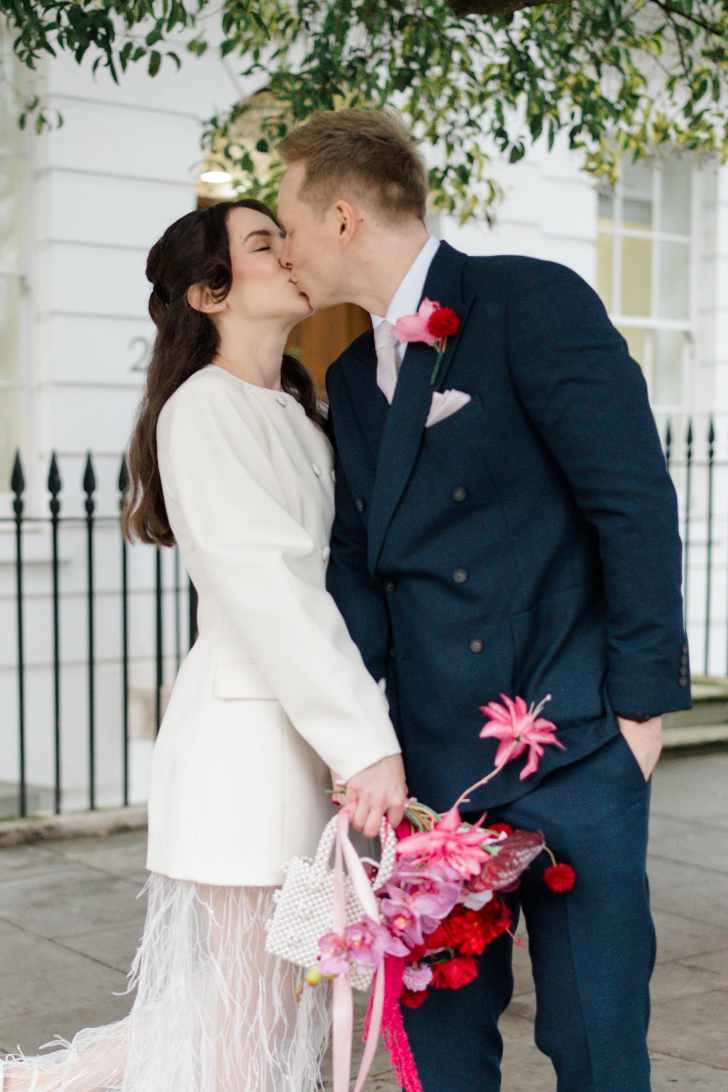 Bride and groom sharing a kiss outdoors, holding vibrant pink and red bridal bouquet.