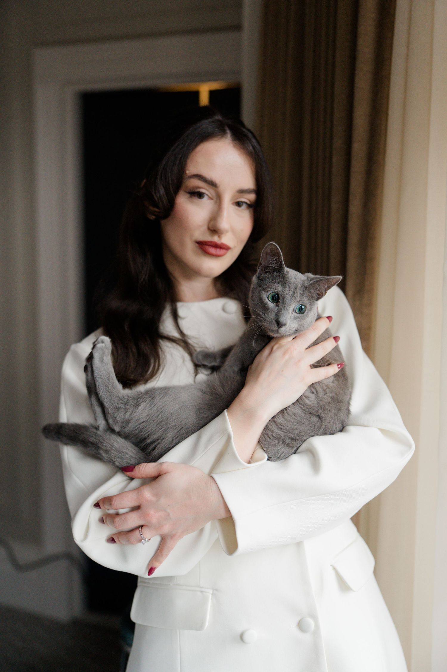 Elegant bride in white double-breasted blazer holding a grey Russian Blue cat indoors.