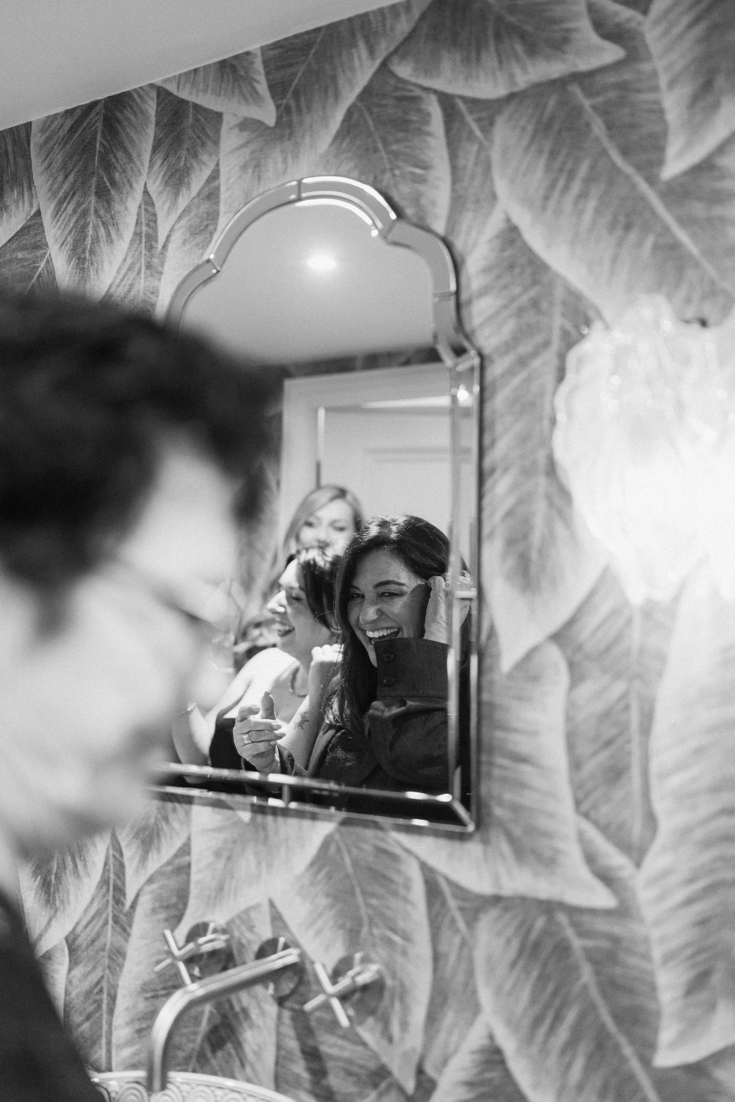 Black and white mirror reflection of a smiling bride getting ready with bridesmaids in a tropical wallpapered bathroom.