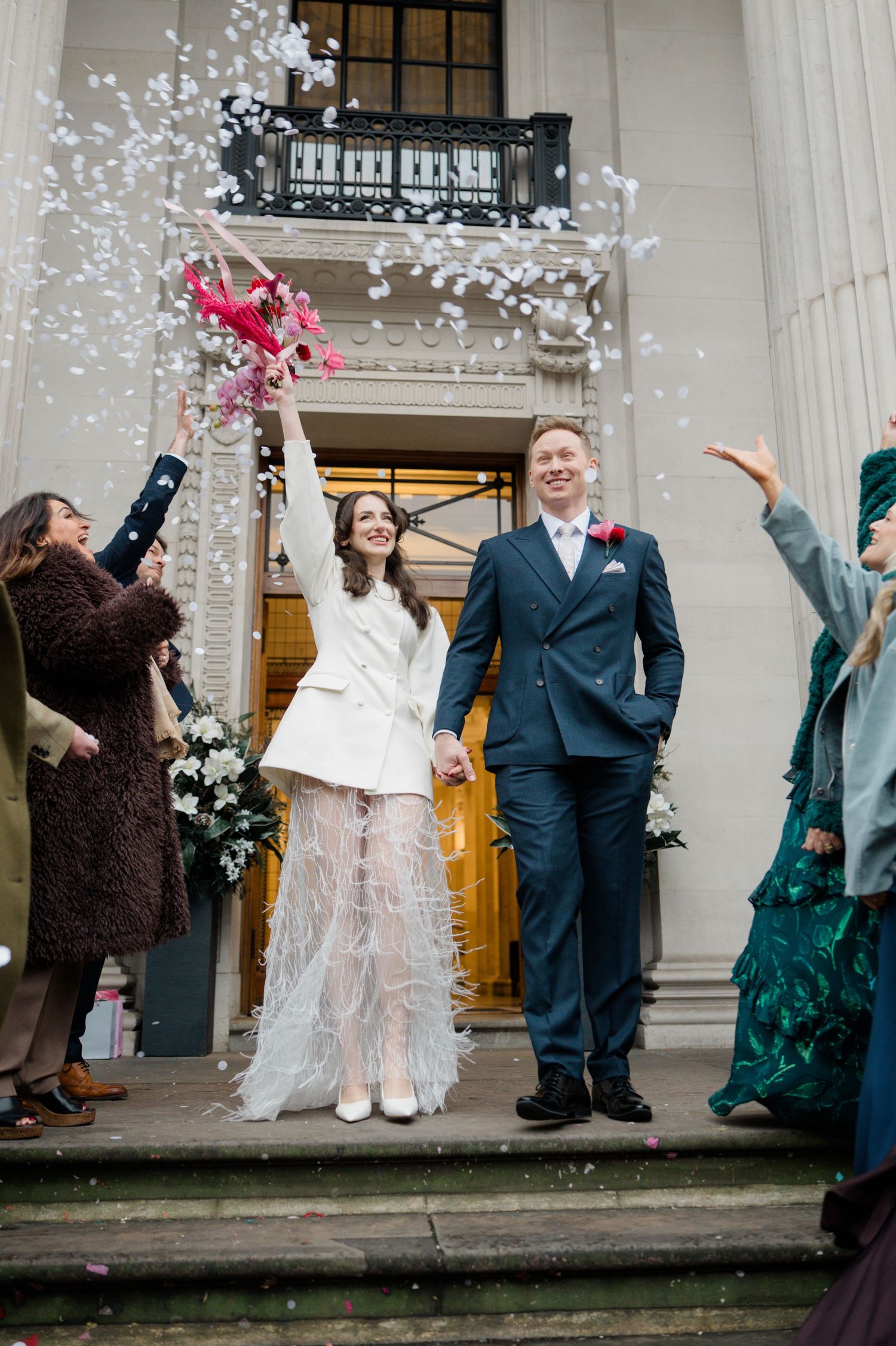 Bride in white feather skirt and groom in navy suit exit venue as guests throw confetti.