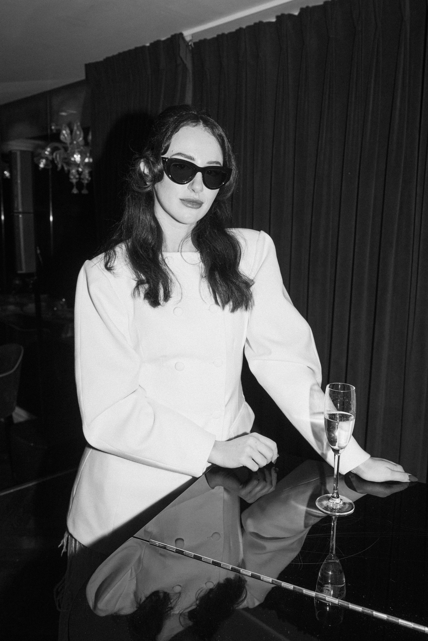 Elegant woman in white blazer with sunglasses holding champagne glass at a bar.