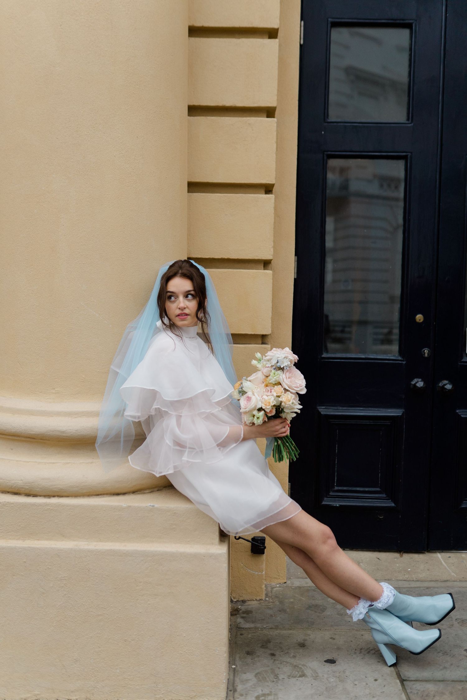 Bride in short white dress, blue veil, and light blue boots sits on steps holding a bouquet of pastel flowers.