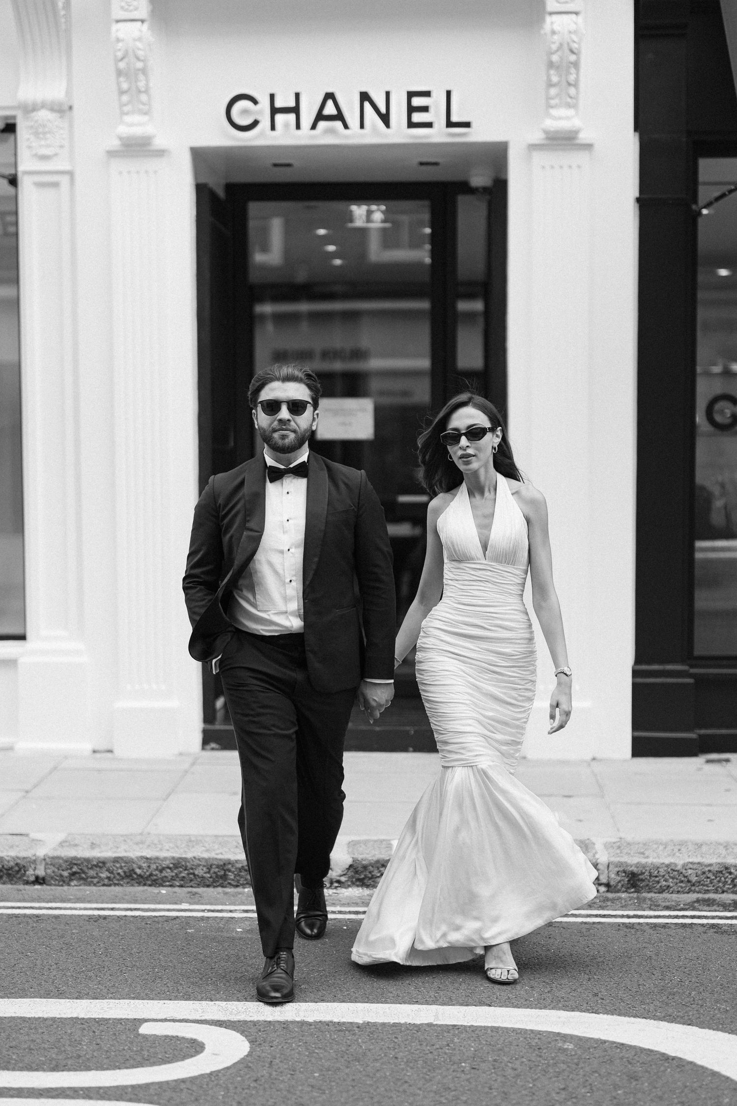 Elegant couple in formal attire walking past a Chanel store, man in tuxedo and woman in white mermaid gown.