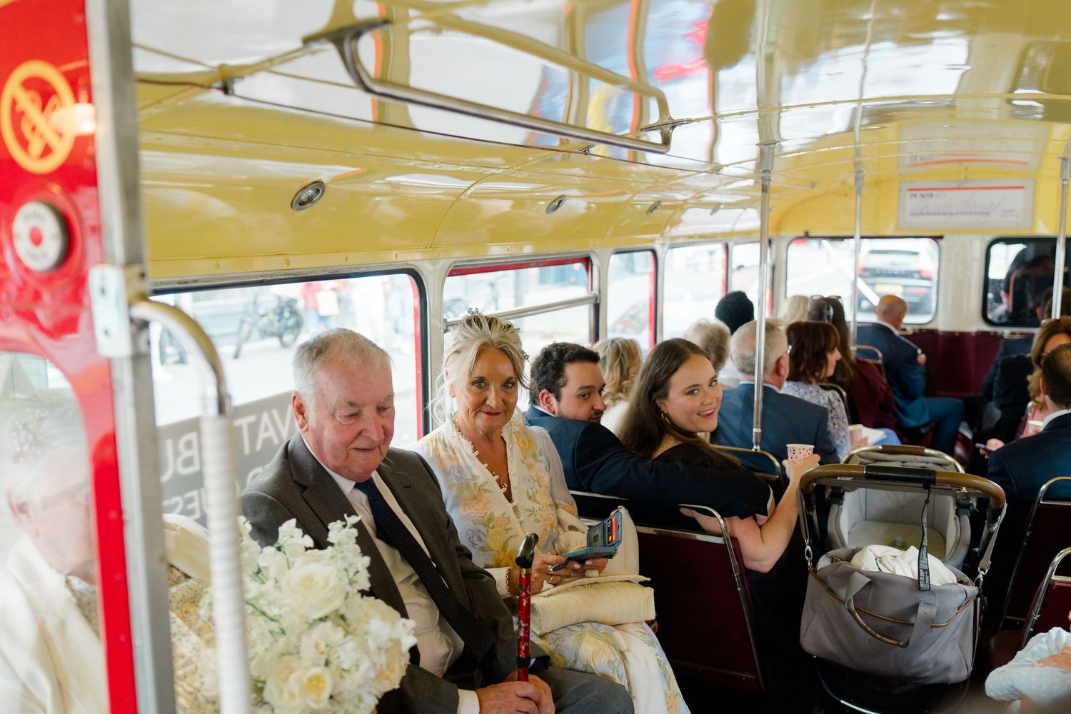 Wedding party guests ride a vintage double-decker bus, bride holding white floral bouquet.