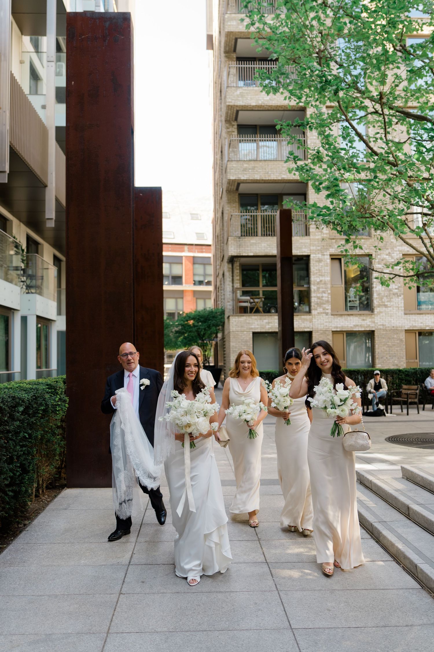 Bride with father and bridesmaids in white gowns walking through a modern urban courtyard with a tall steel sculpture.