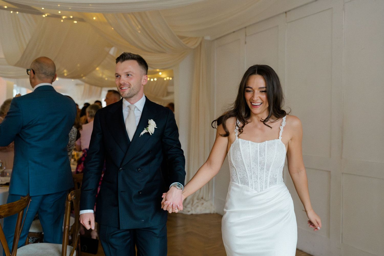 Bride in white corset gown and groom in navy suit enter wedding reception holding hands and smiling.