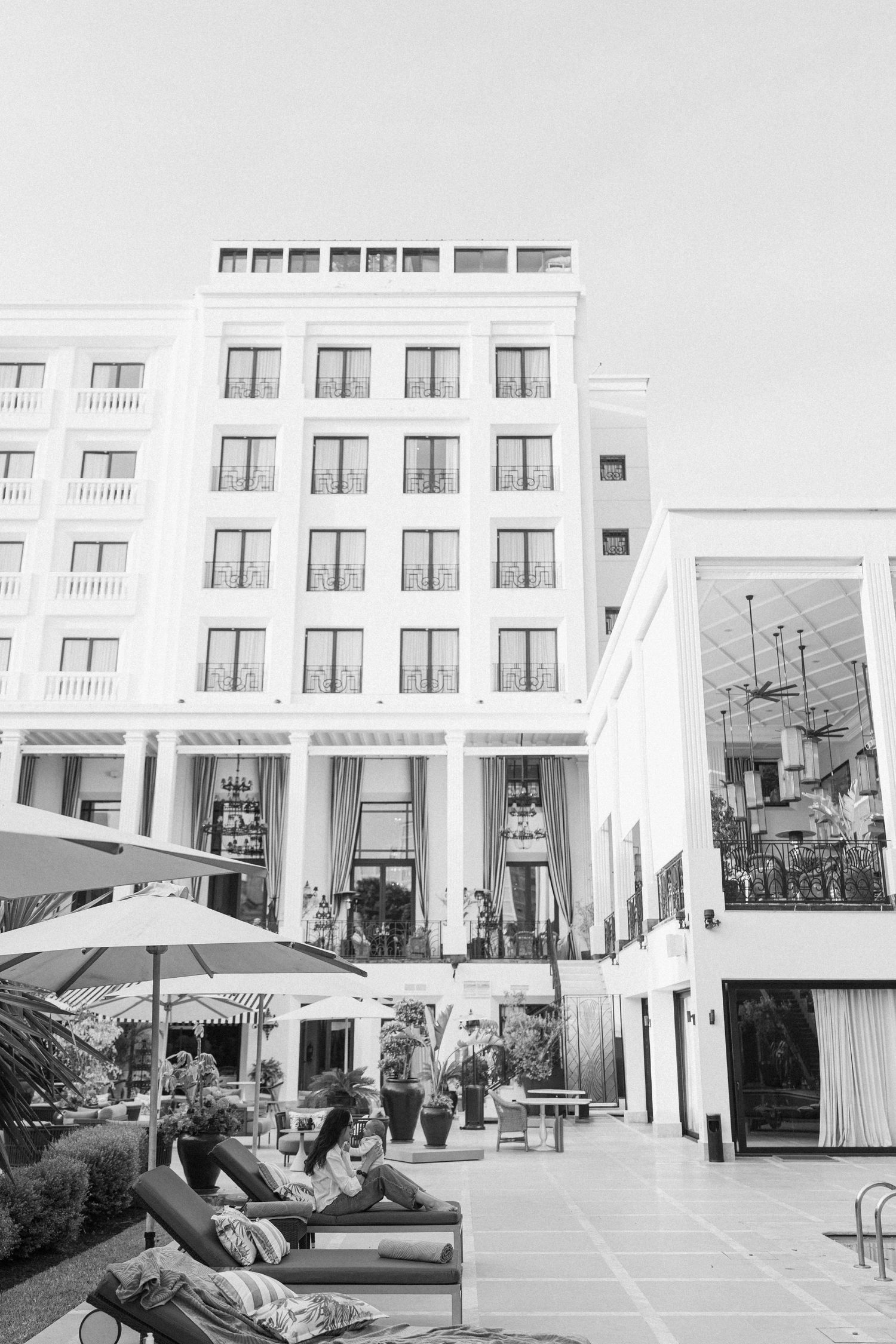 Black and white photo of guests relaxing on lounge chairs by a hotel pool with elegant white building facade.