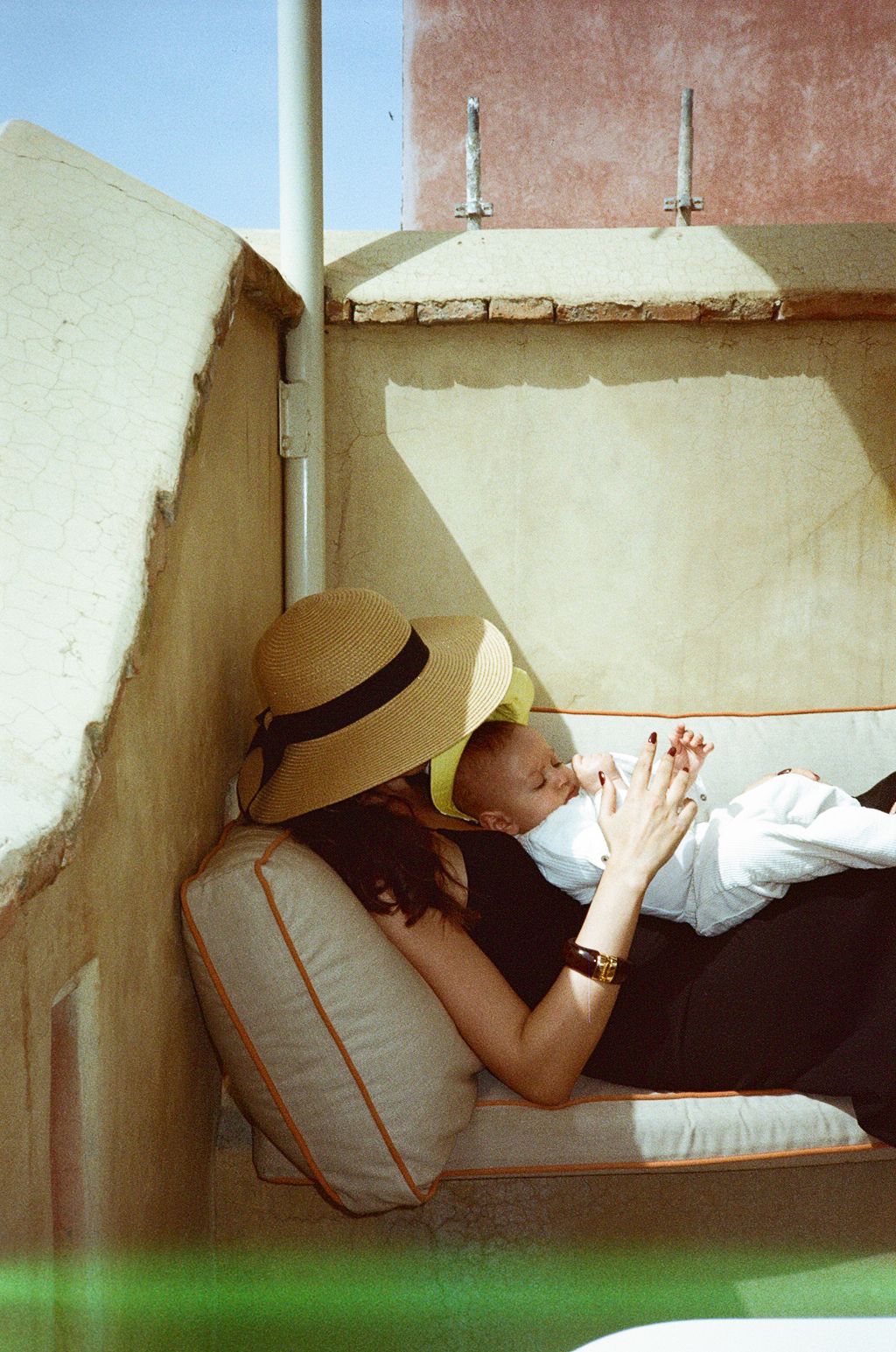 Mother in straw hat breastfeeding newborn baby while reclining on cushioned outdoor lounger in sunny Mediterranean setting.