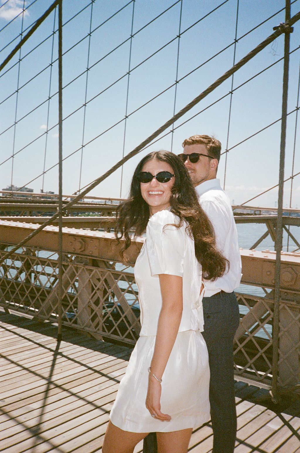 Smiling couple in sunglasses posing on Brooklyn Bridge walkway on a sunny day.