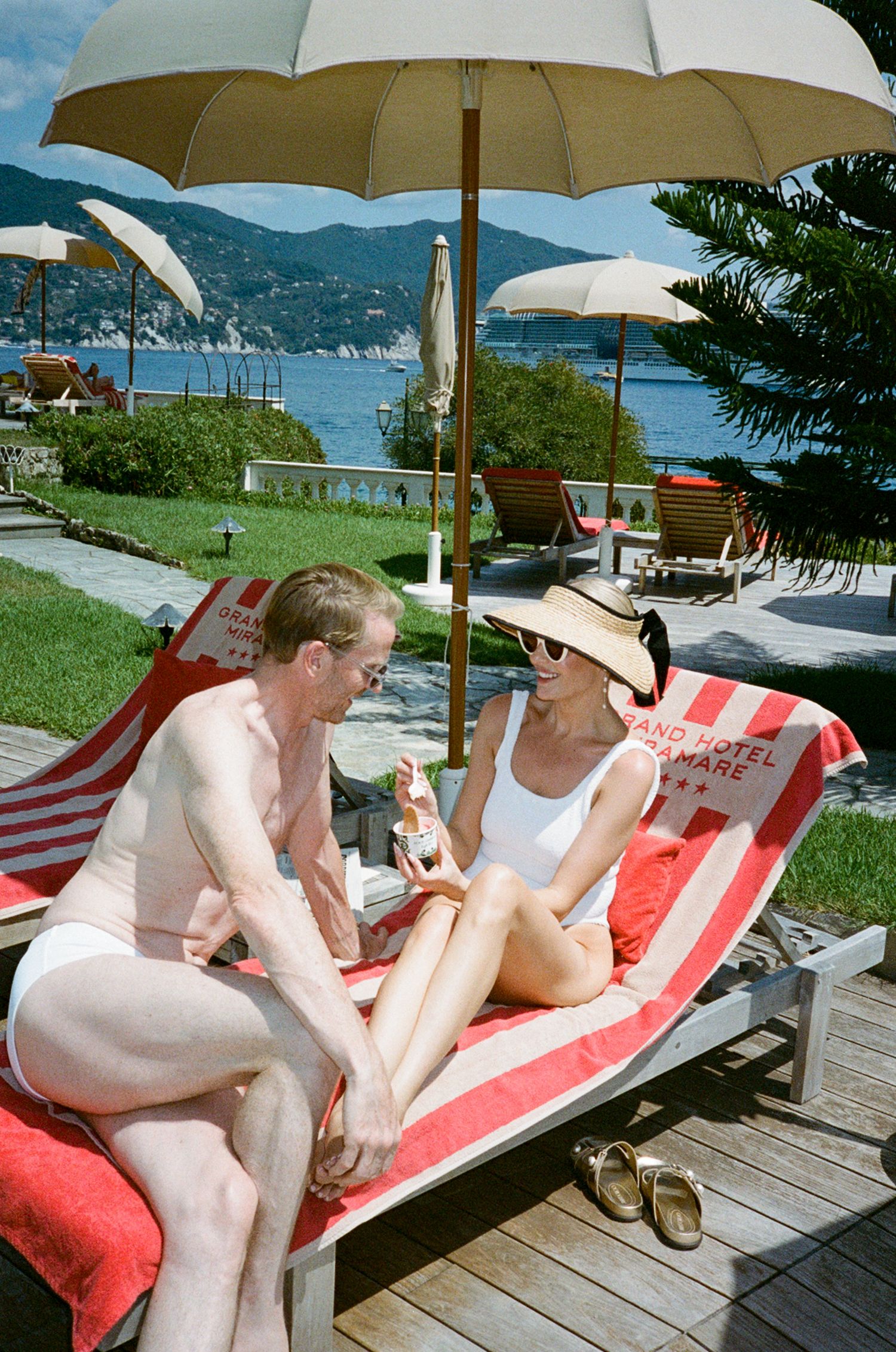 Couple relaxing on red striped lounge chairs under beach umbrella at Grand Hotel sul Mare by Italian lake.