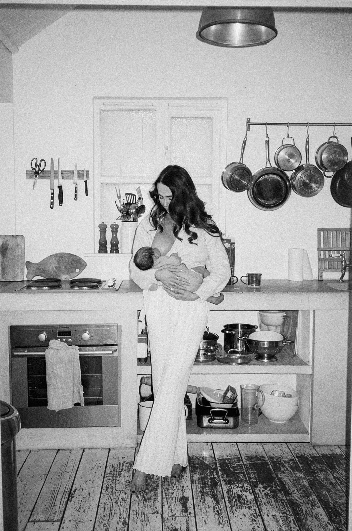 Black and white photo of a woman in white outfit holding a newborn baby in a rustic kitchen with hanging pots.