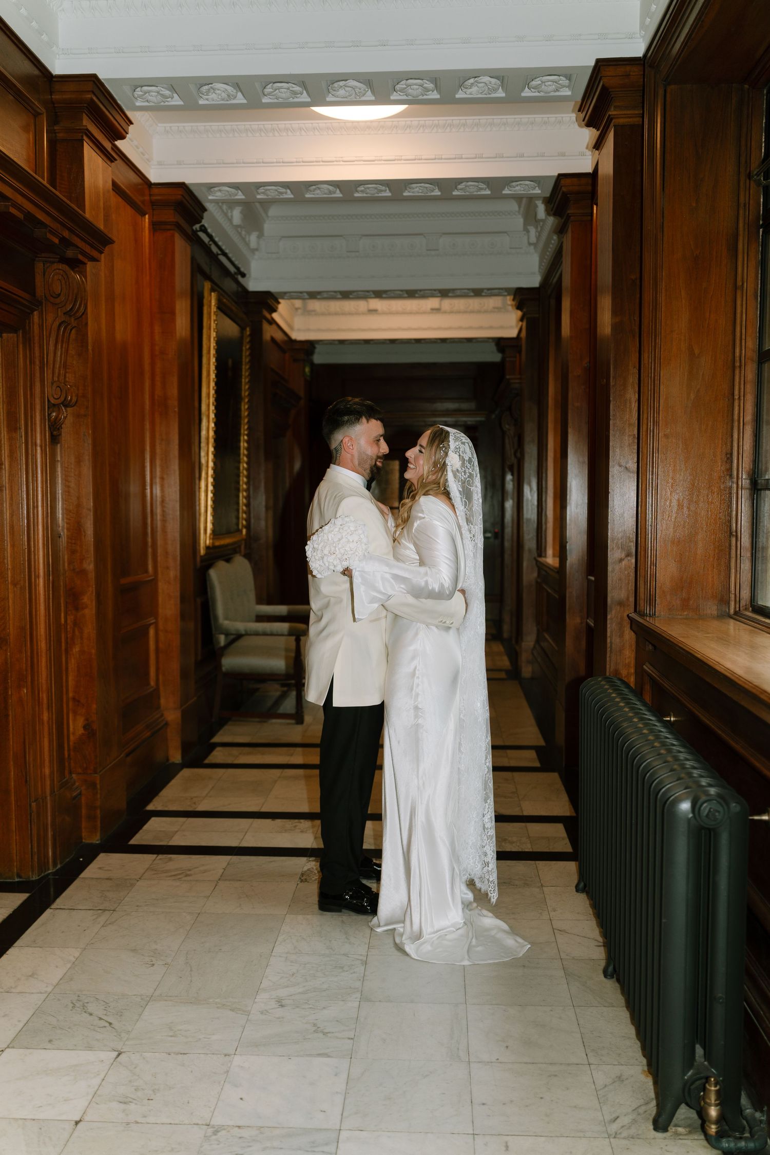 A couple in white wedding attire shares an intimate moment in an elegant wood-paneled hallway.