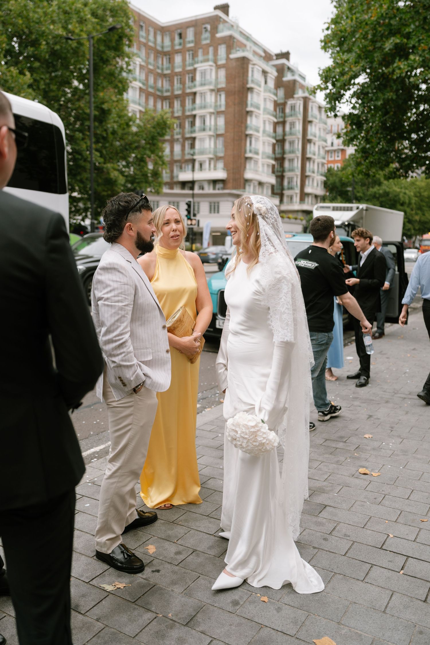 Wedding guests in formal attire gather outside a brick building on a paved street.