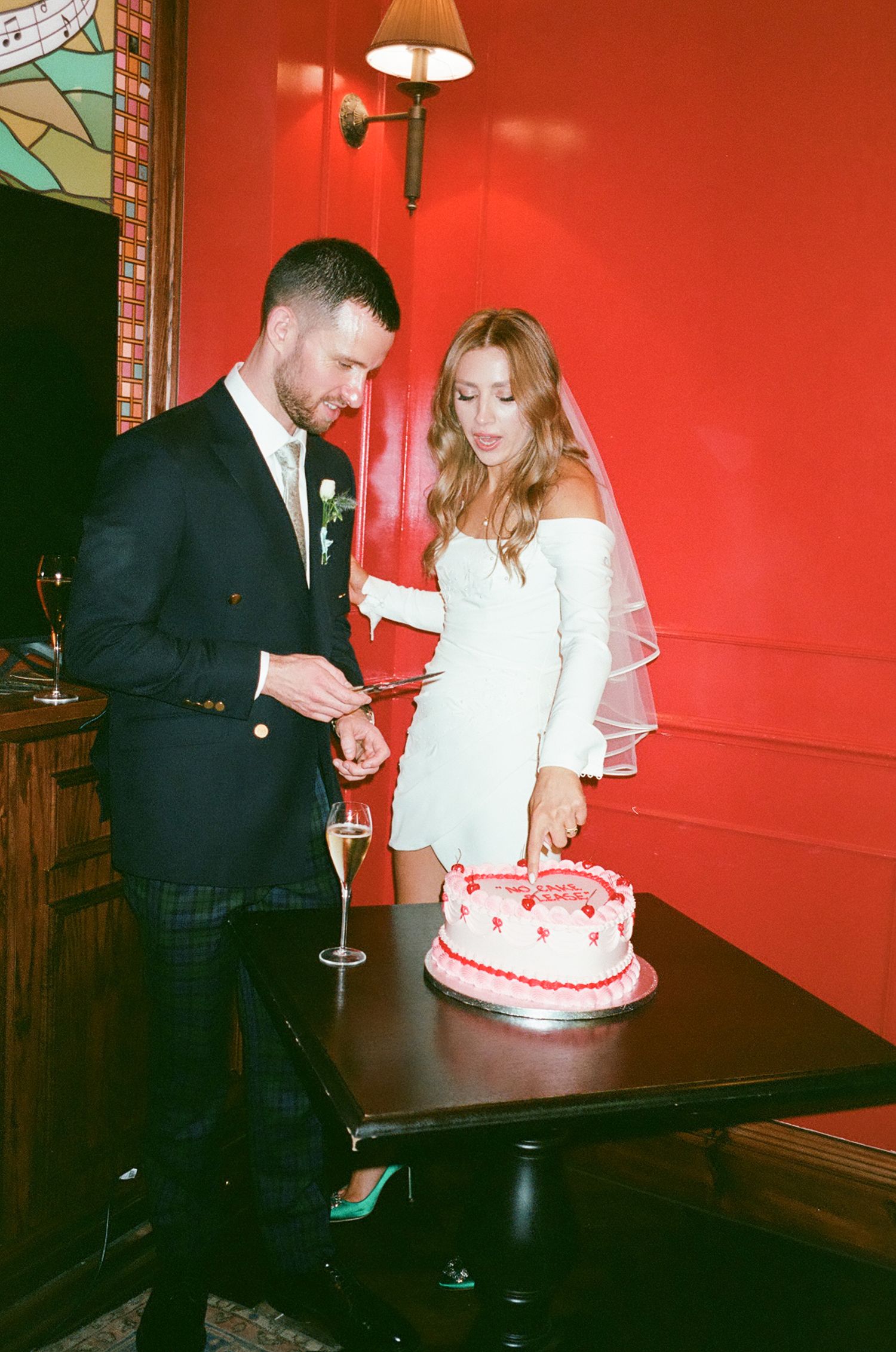 A couple in formal attire cuts into a white and red wedding cake in a room with red walls.