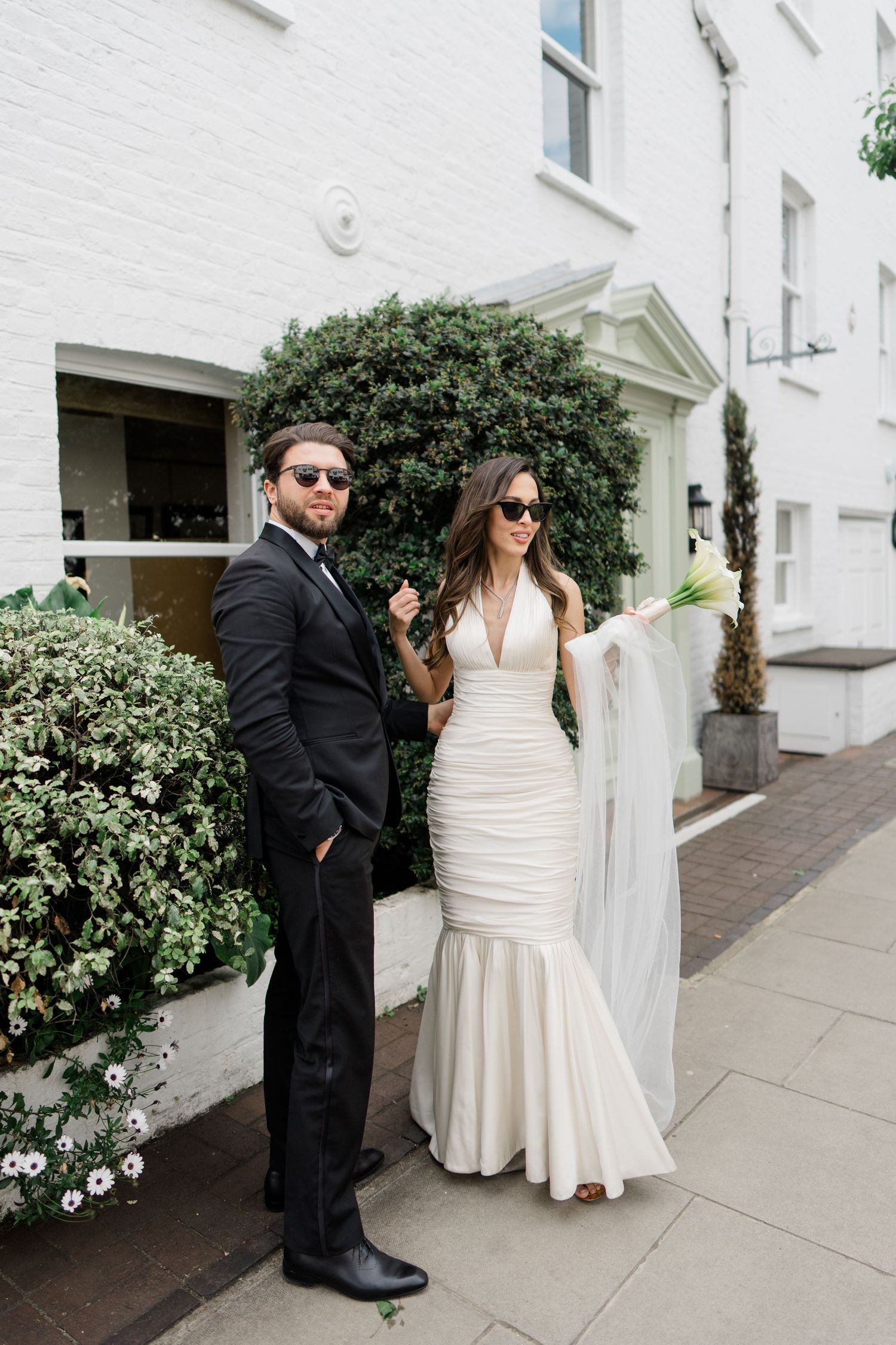 A couple poses against a white building exterior with decorative shrubbery.