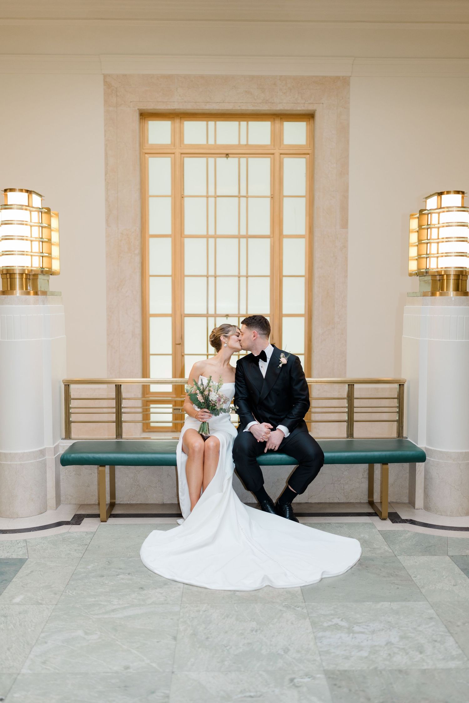 Elegant wedding couple sits on green bench in front of large window with decorative wall sconces in formal venue.