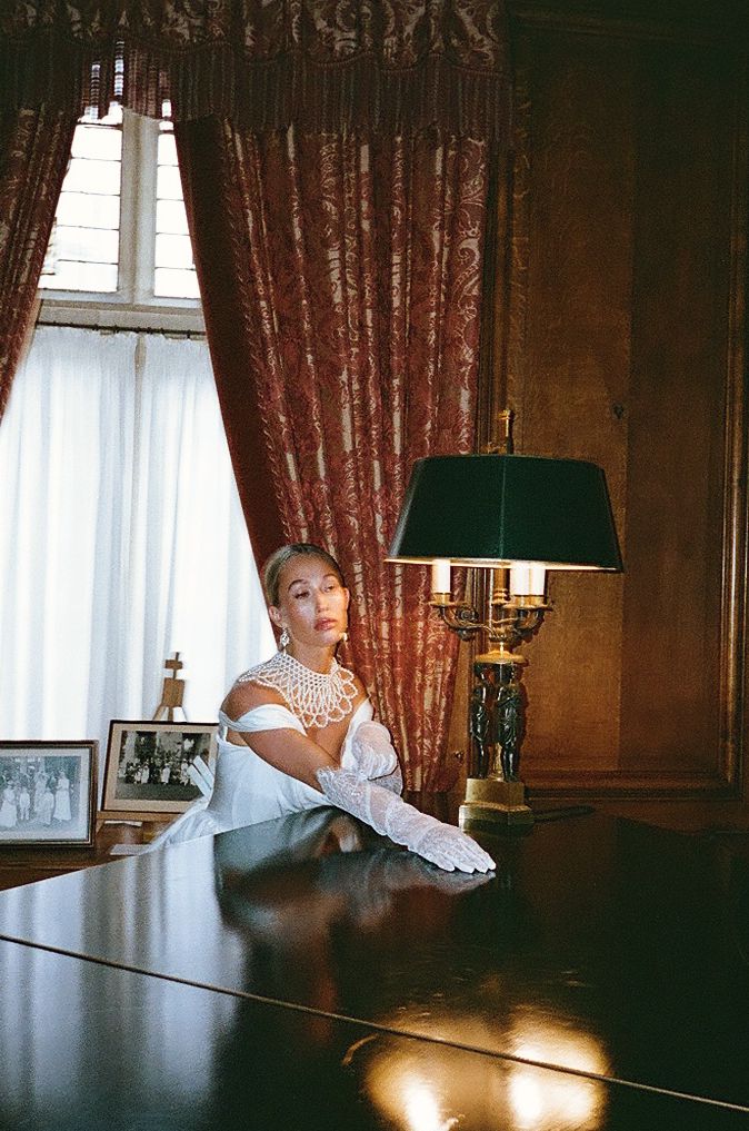 A figure sits at a long dark wooden table in an elegant room with red curtains and natural light streaming through windows.