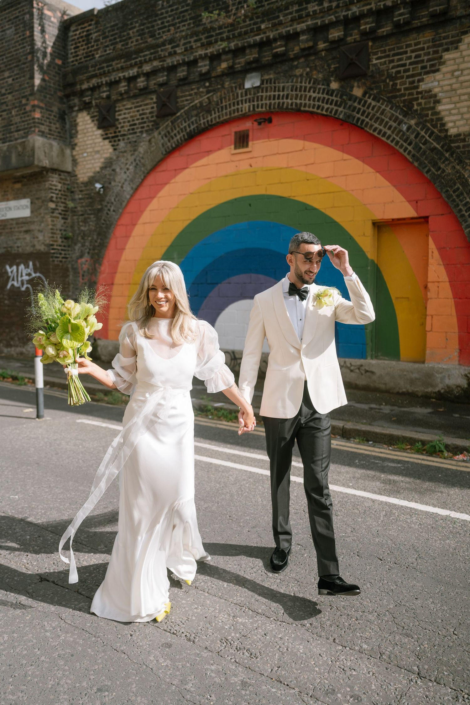 A couple poses in front of a vibrant rainbow-painted brick archway with the bride holding a bouquet.