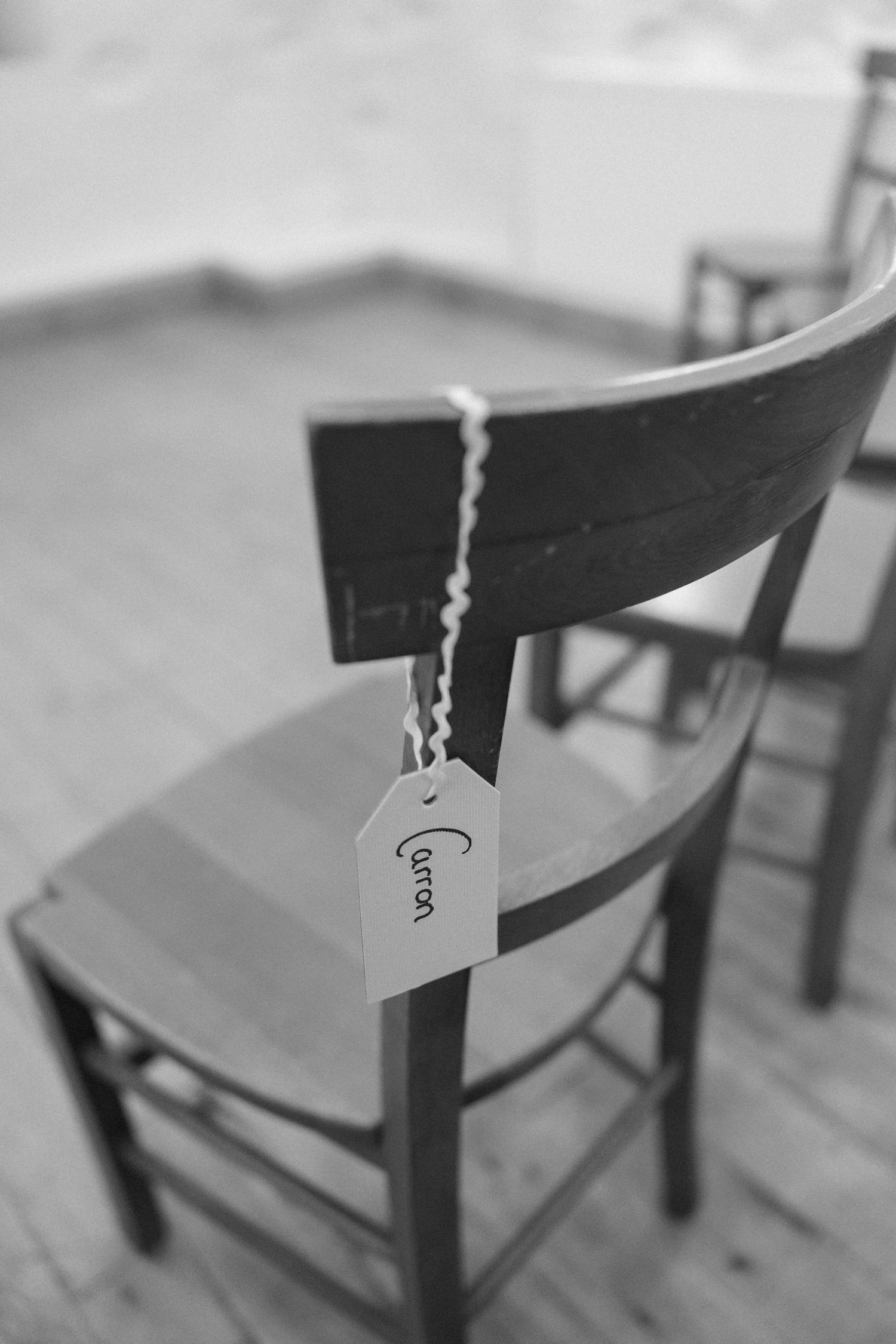 Black and white photo of a wooden chair with a name tag attached.