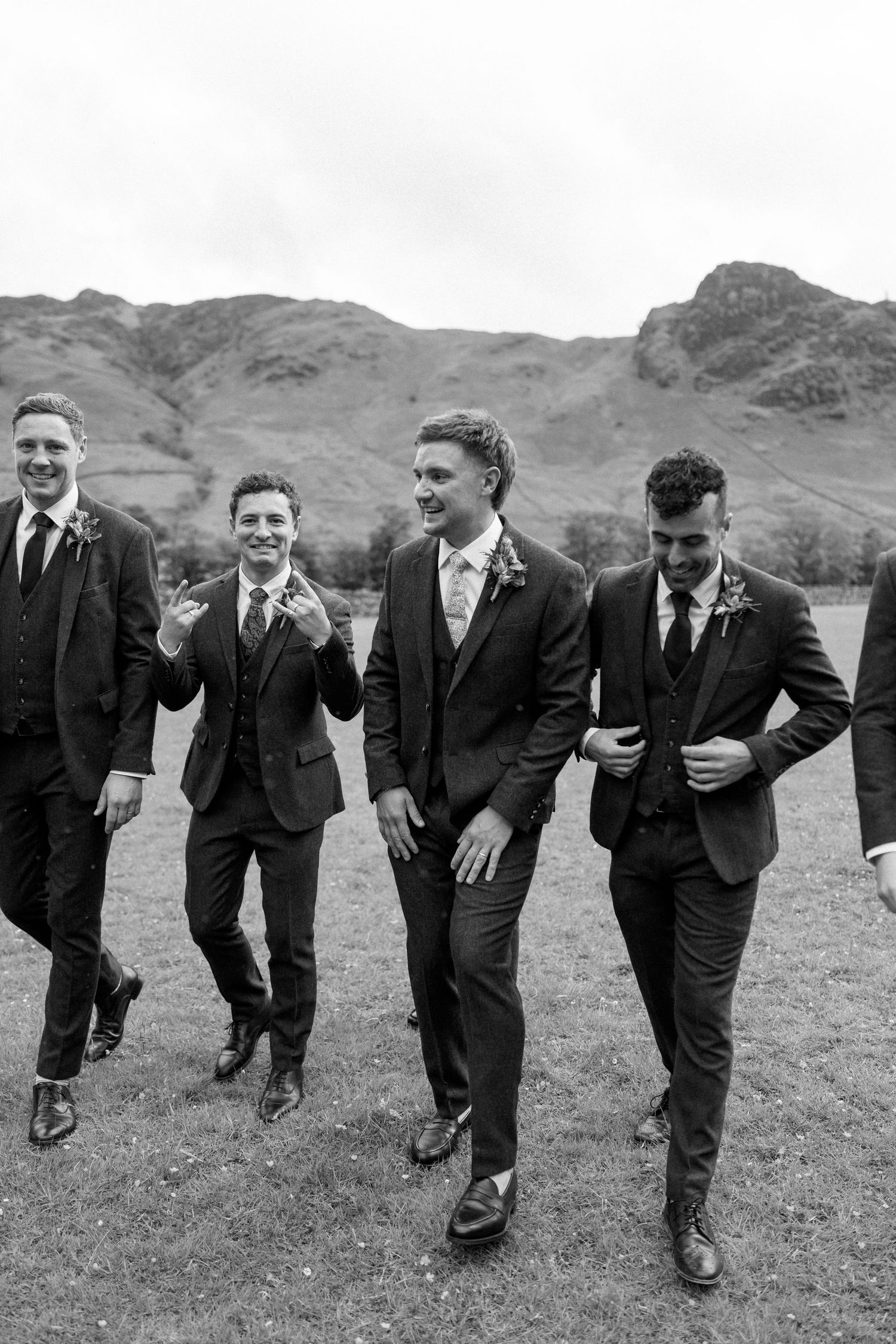 Groomsmen in dark suits pose playfully on a grassy hill with mountains behind them.