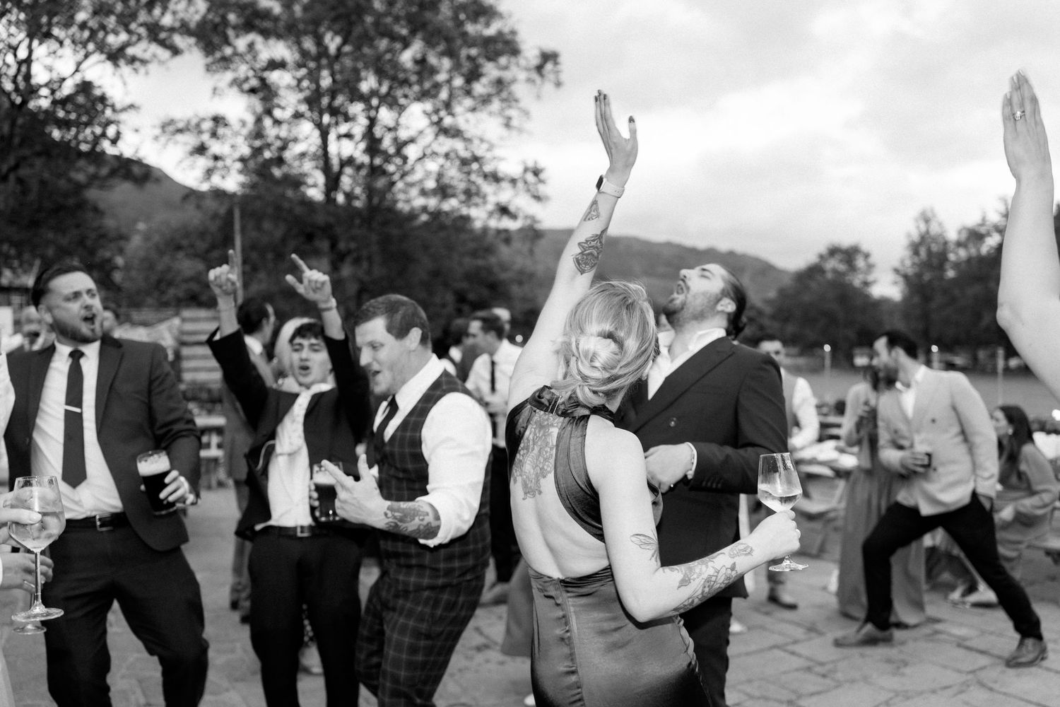 Wedding guests dance in a line outdoors with mountains and trees in the background.