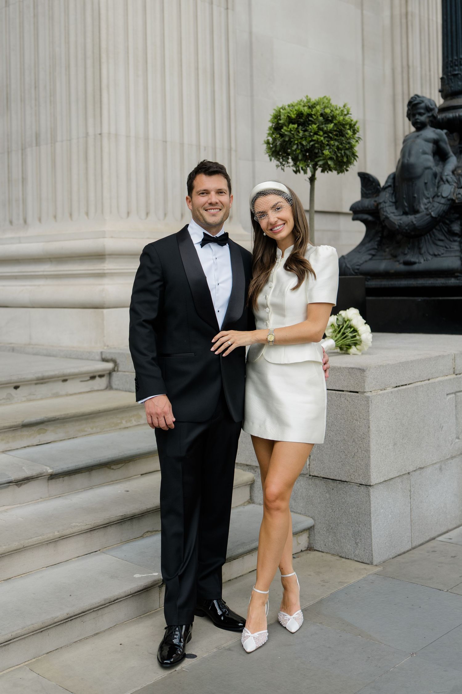 Couple in wedding attire on stone steps, groom in black tuxedo, bride in white mini dress holding bouquet.