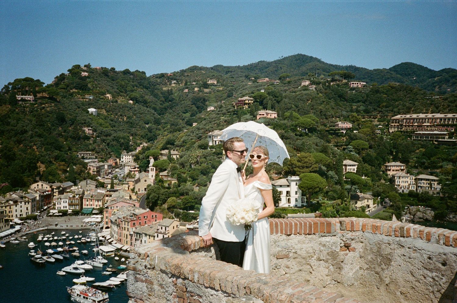 Wedding couple kissing under white umbrella overlooking Portofino harbor and colorful hillside village in Italy.