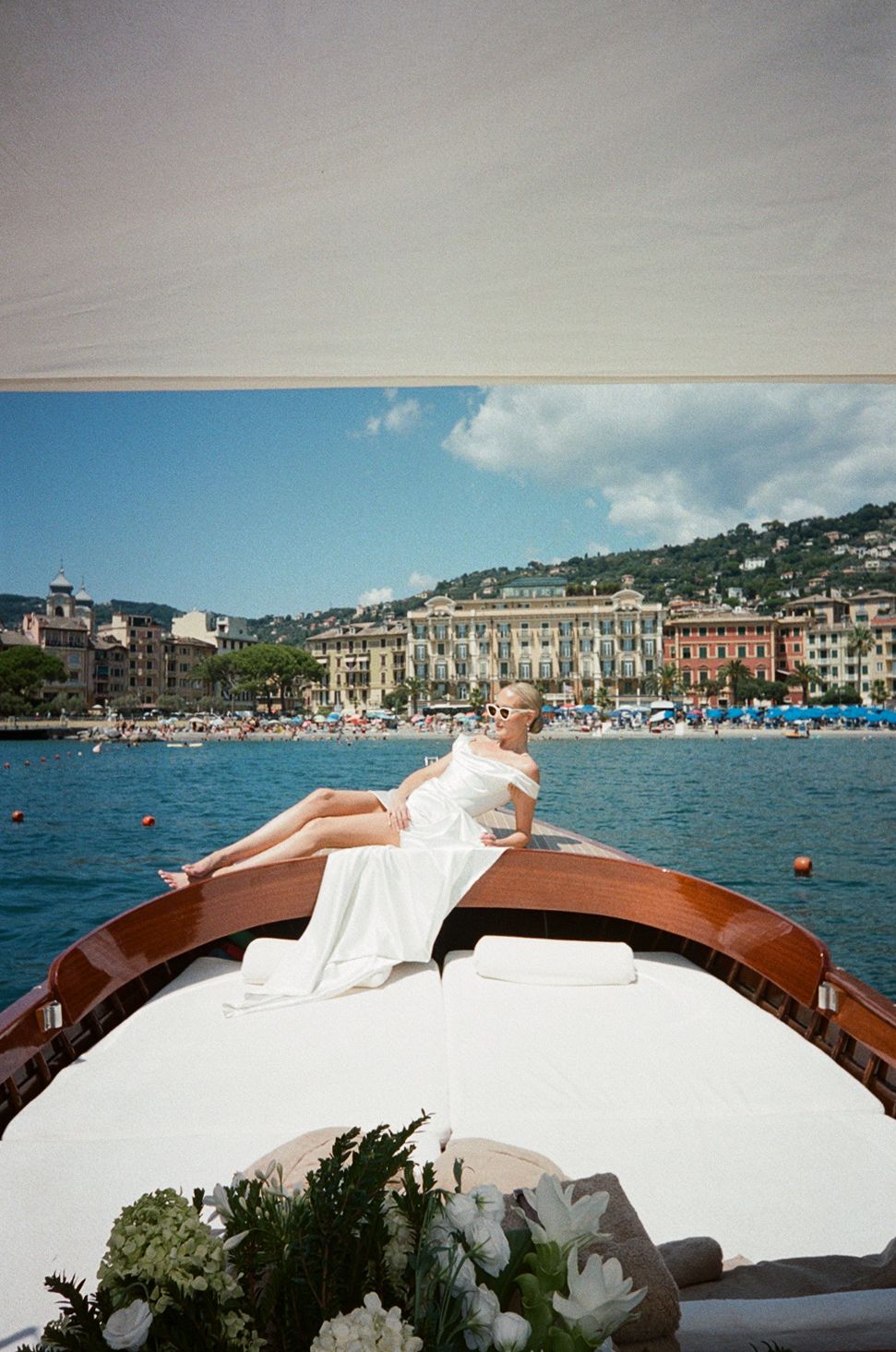 Bride in white off-shoulder gown lounging on bow of wooden boat in Italian harbor.