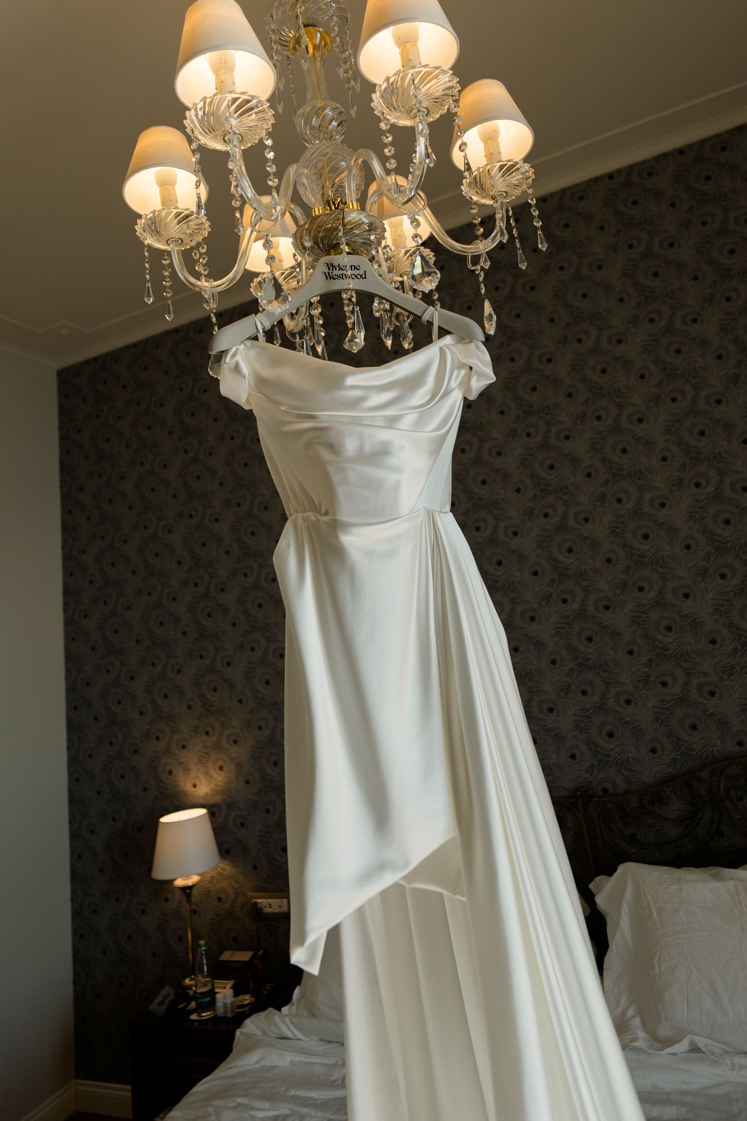 Elegant white off-shoulder wedding dress hanging from a crystal chandelier in a luxurious hotel bedroom.