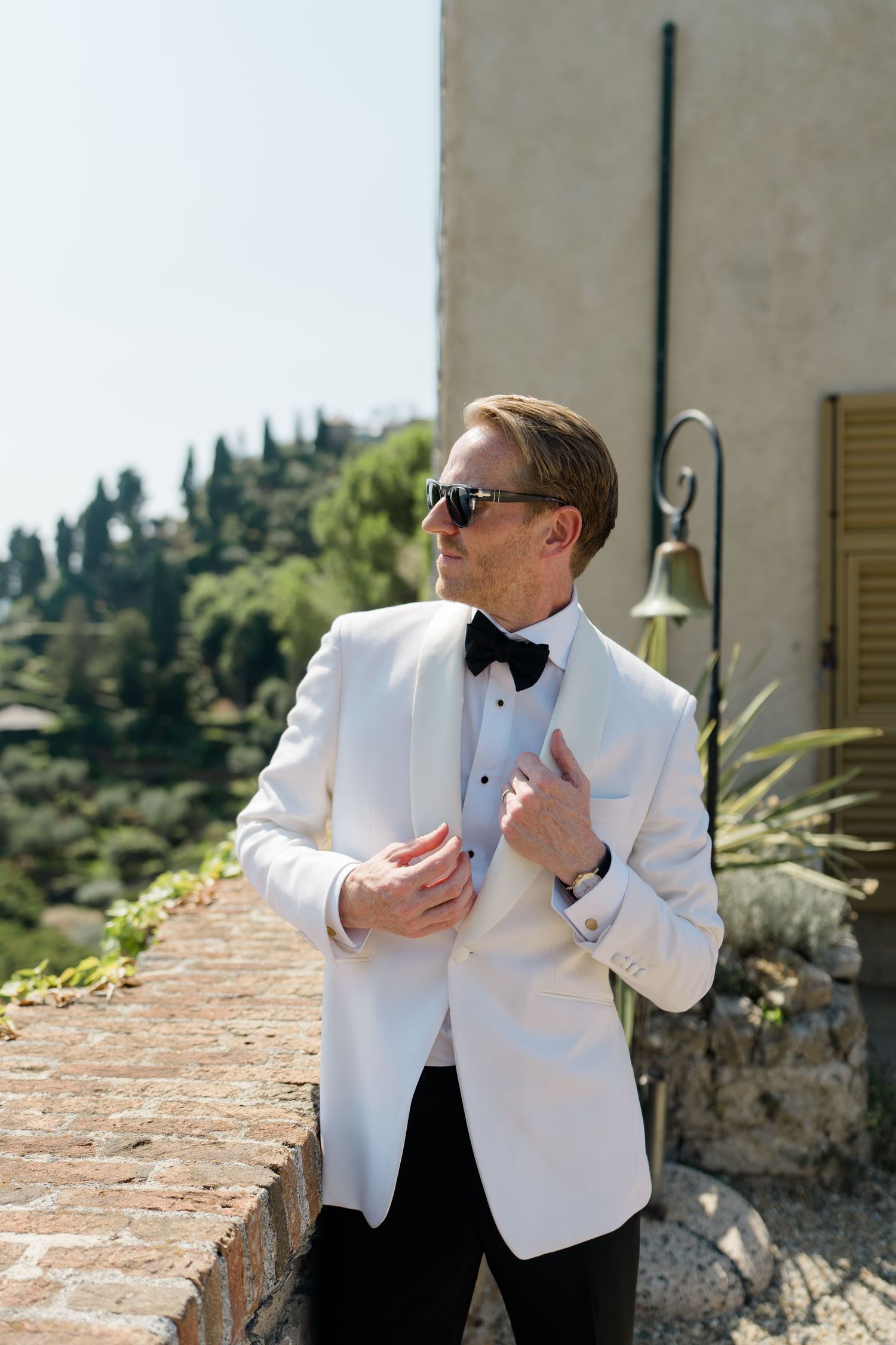 Groom in white tuxedo jacket and black bow tie adjusts suit outdoors with scenic view.