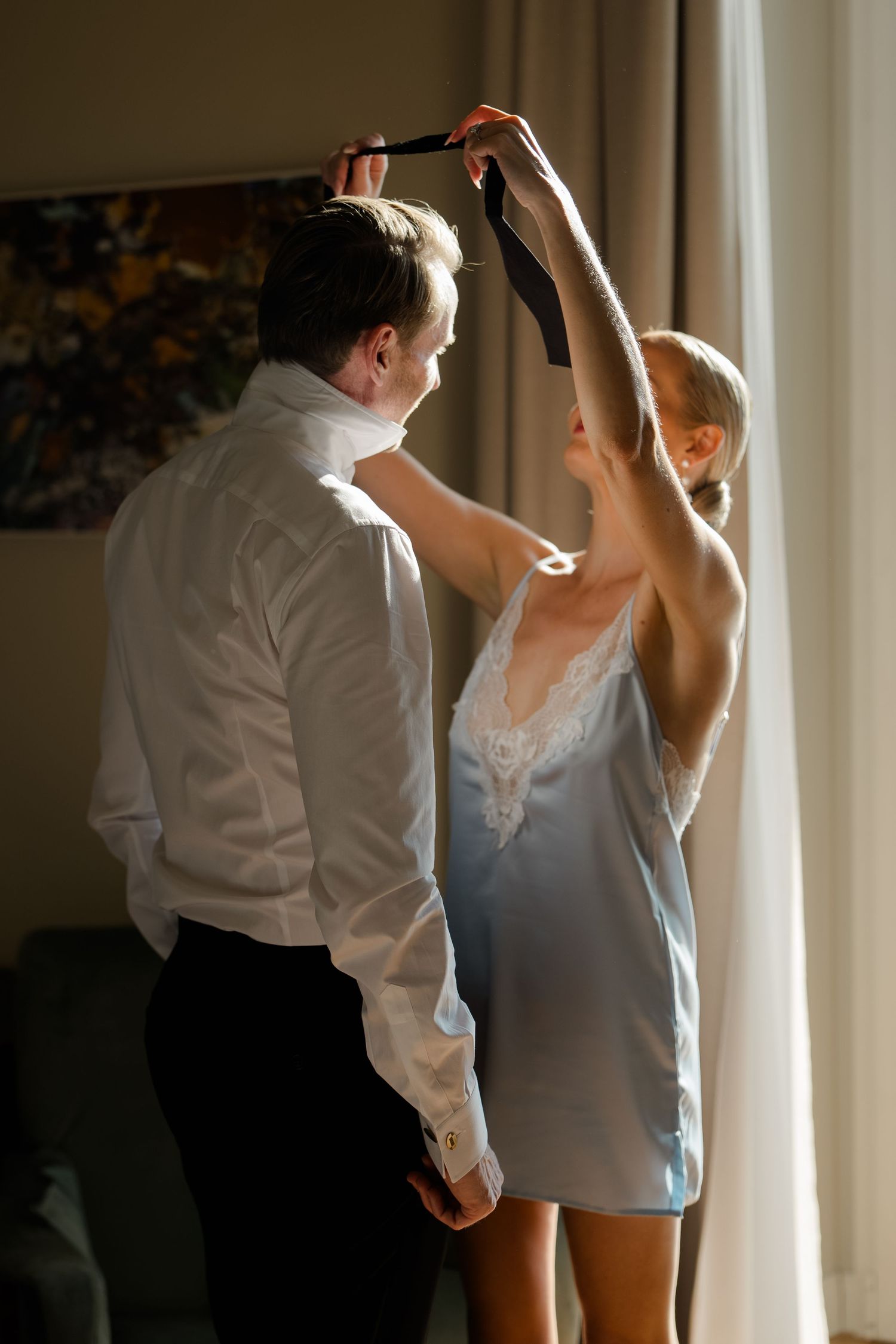Woman in blue slip dress helping a man in white shirt put on a black tie in a sunlit room.