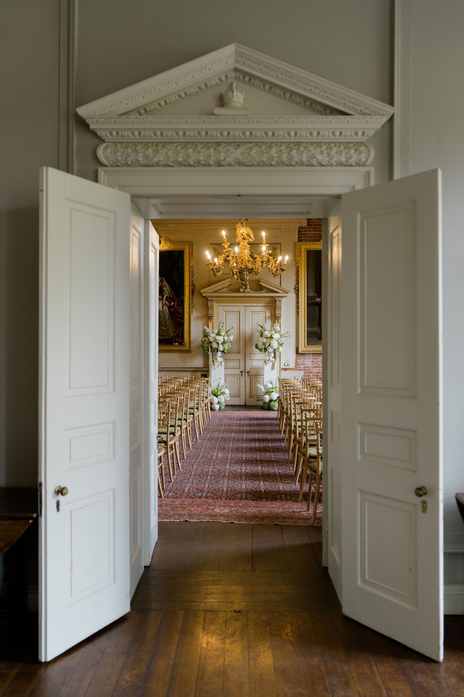Elegant wedding ceremony setup viewed through open white double doors with chandelier and floral arrangements.