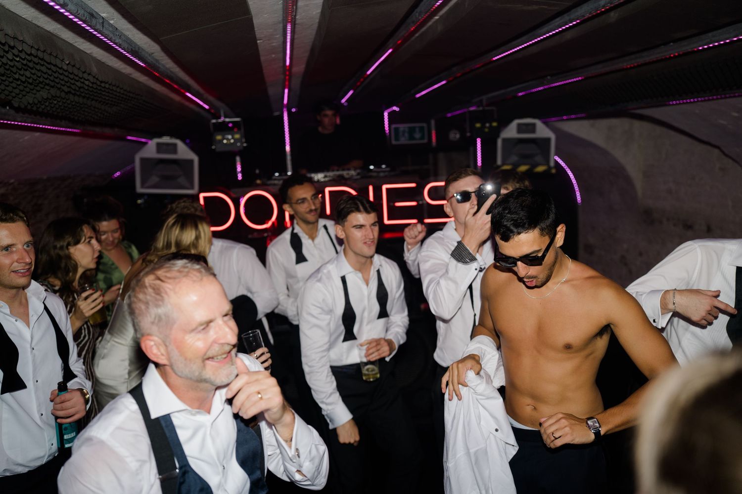 Energetic wedding after-party with guests dancing in white shirts under purple neon lights.