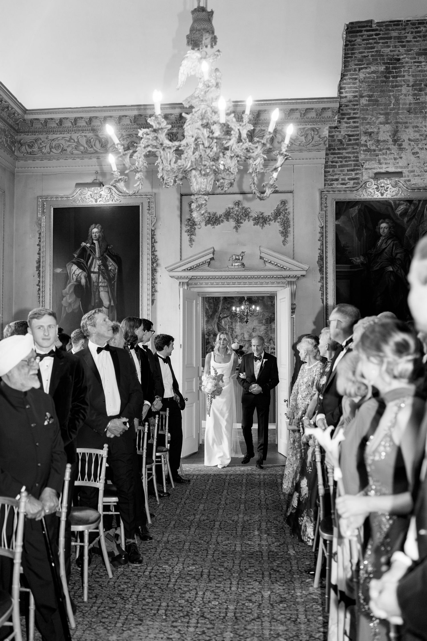 Black and white photo of bride walking down aisle with father in elegant chandelier ballroom.
