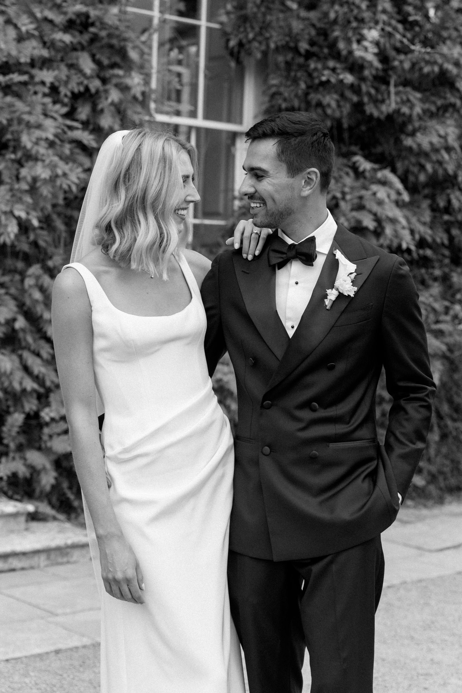 Bride and groom smile at each other outdoors, she in sleek white dress, he in black tuxedo with boutonniere.