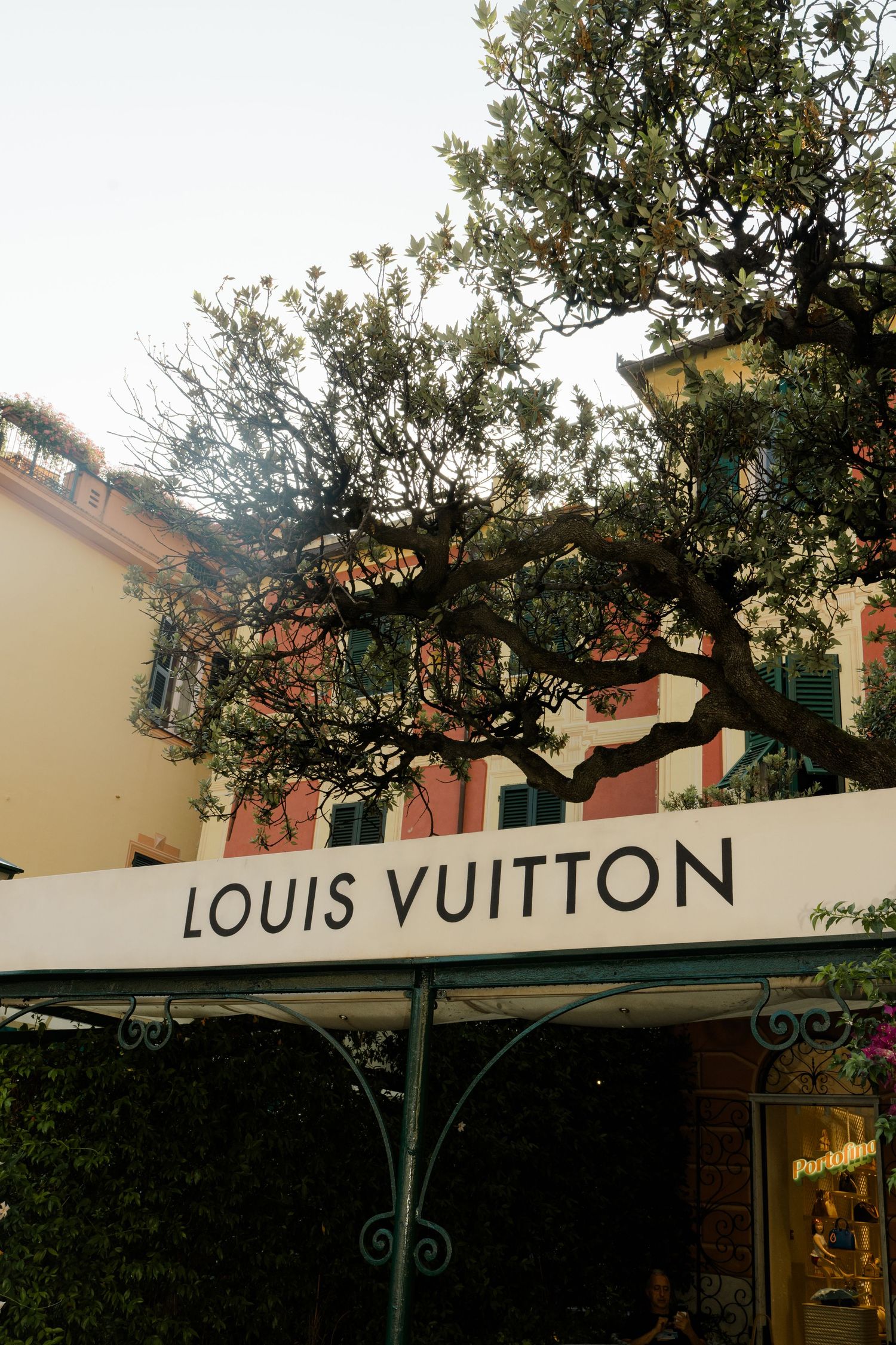 Louis Vuitton luxury boutique storefront in Portofino Italy framed by olive tree branches and colorful buildings.