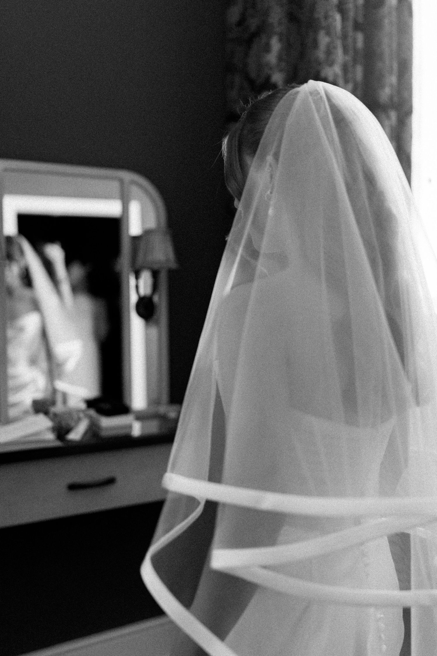 Bride wearing a flowing white wedding veil standing before a dressing table mirror.