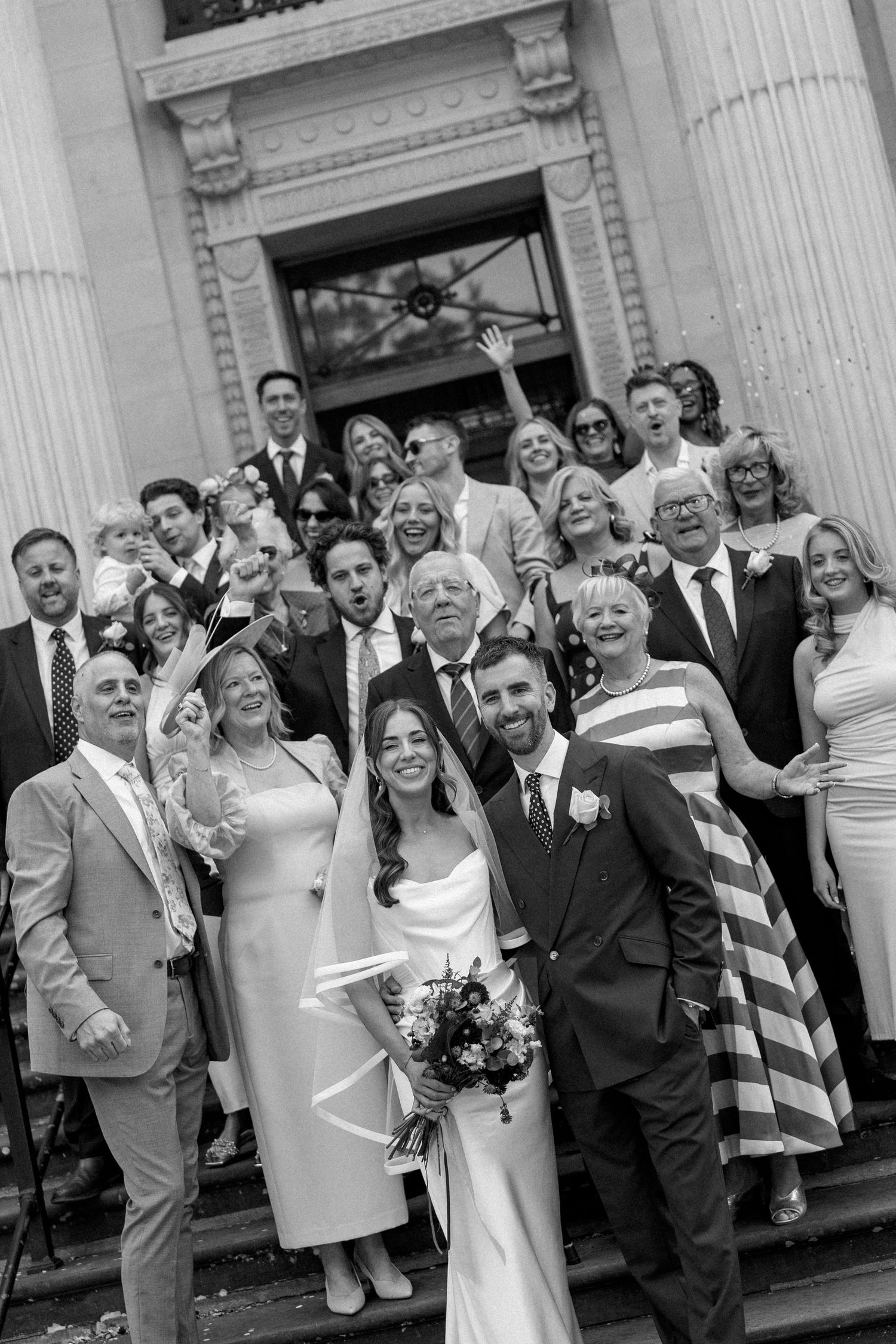 Black and white group wedding photo of bride, groom, and guests celebrating on grand stone steps.