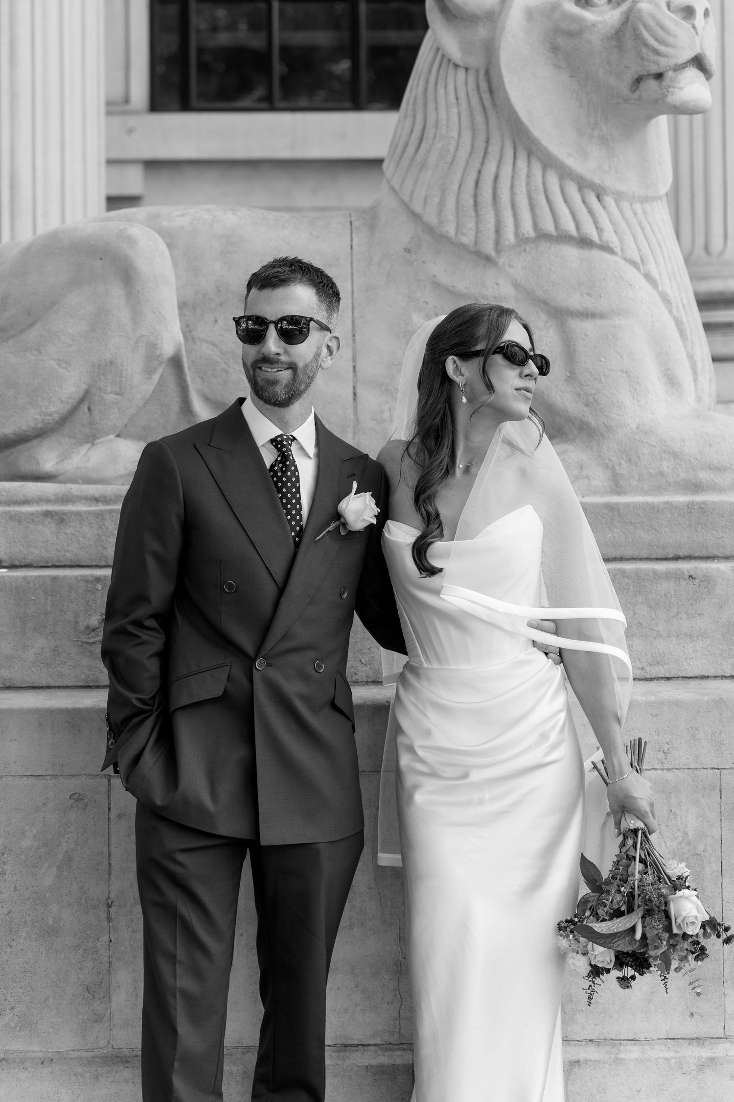 Elegant bride and groom wearing sunglasses pose beside a stone lion sculpture after their ceremony.