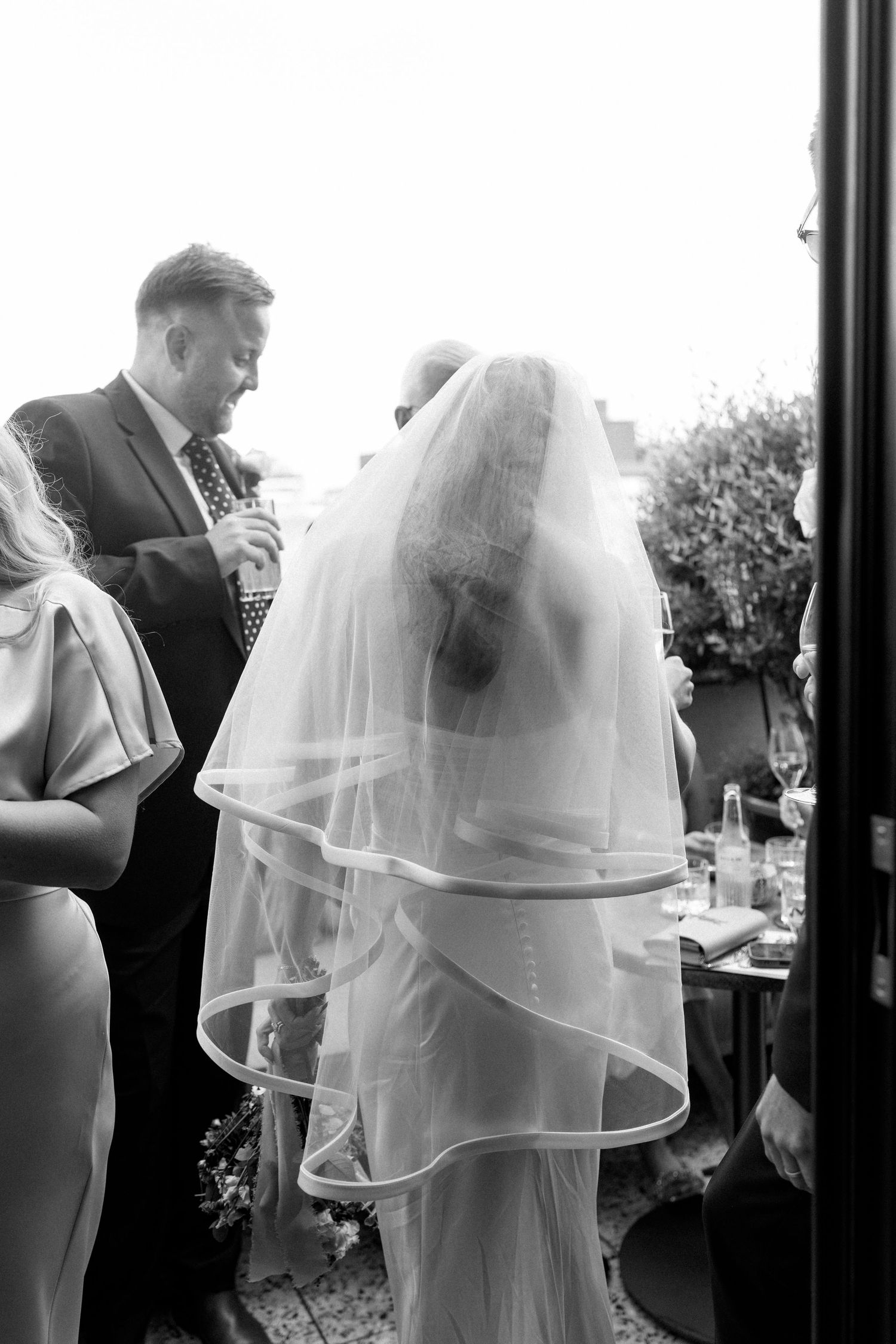 Black and white photo of bride in layered veil standing with wedding guests at outdoor reception.