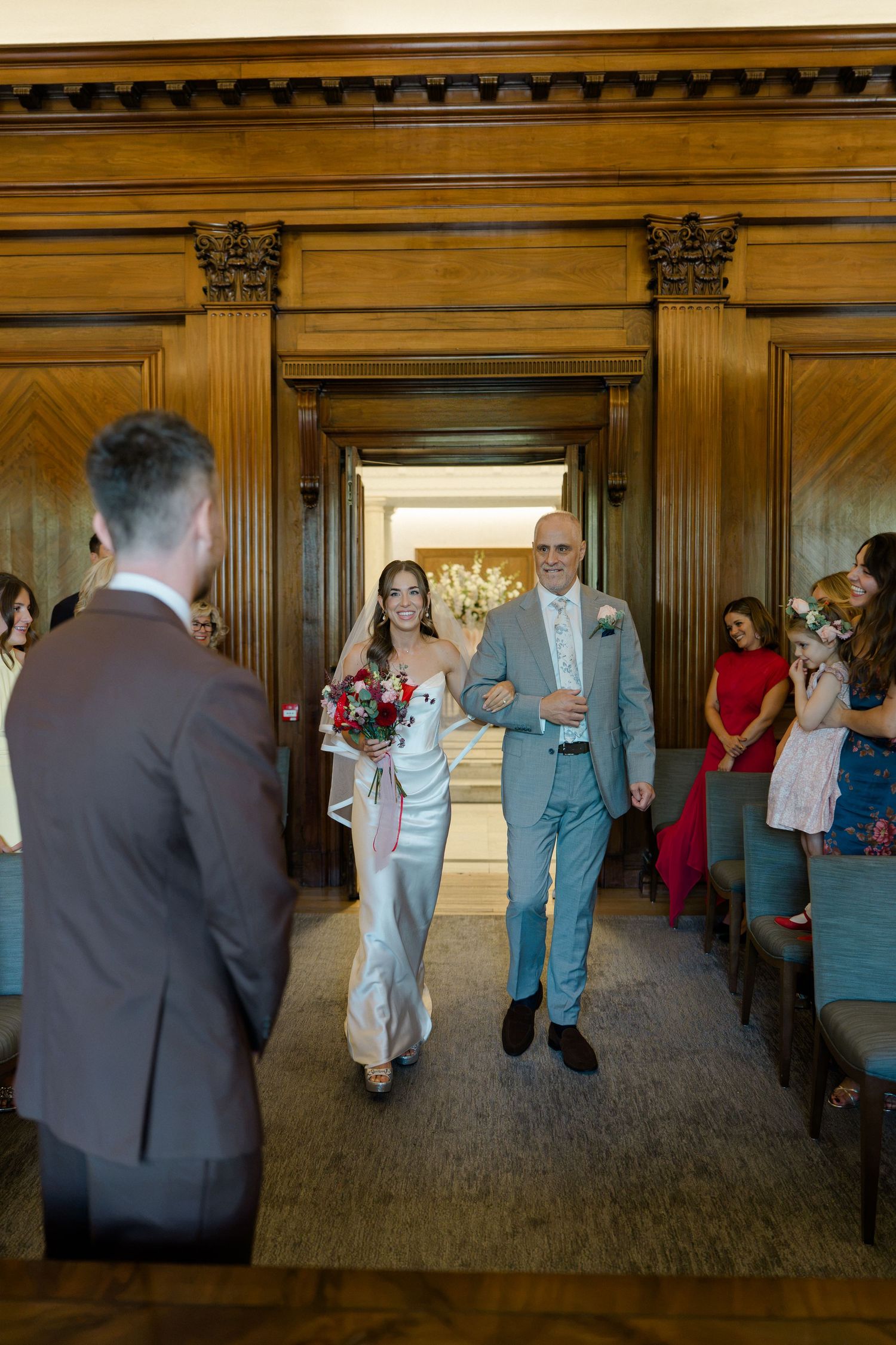 Bride walks down the aisle with her father in an elegant wood-paneled wedding ceremony venue.