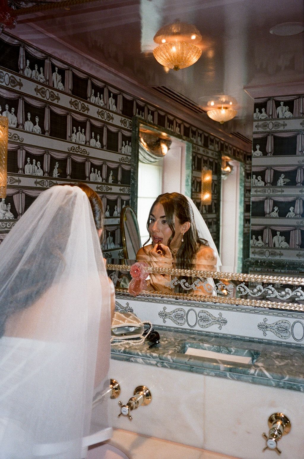 Bride in veil touches up lipstick in ornate gold and marble bathroom mirror.