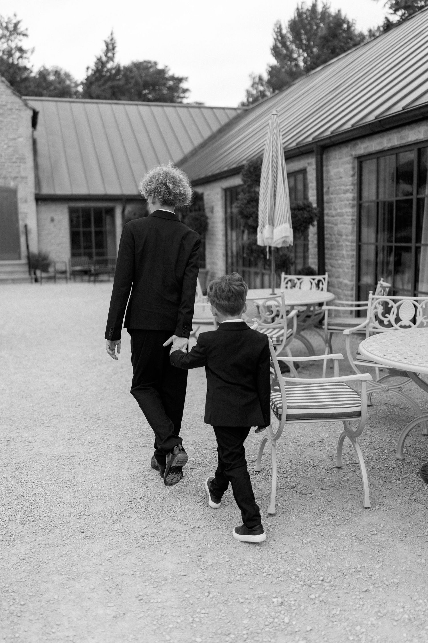 Two boys in black suits walking hand in hand across gravel courtyard at wedding venue.