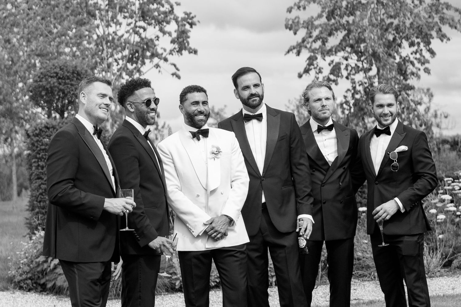 Black and white photo of groom in white jacket posing with five groomsmen in black tuxedos.