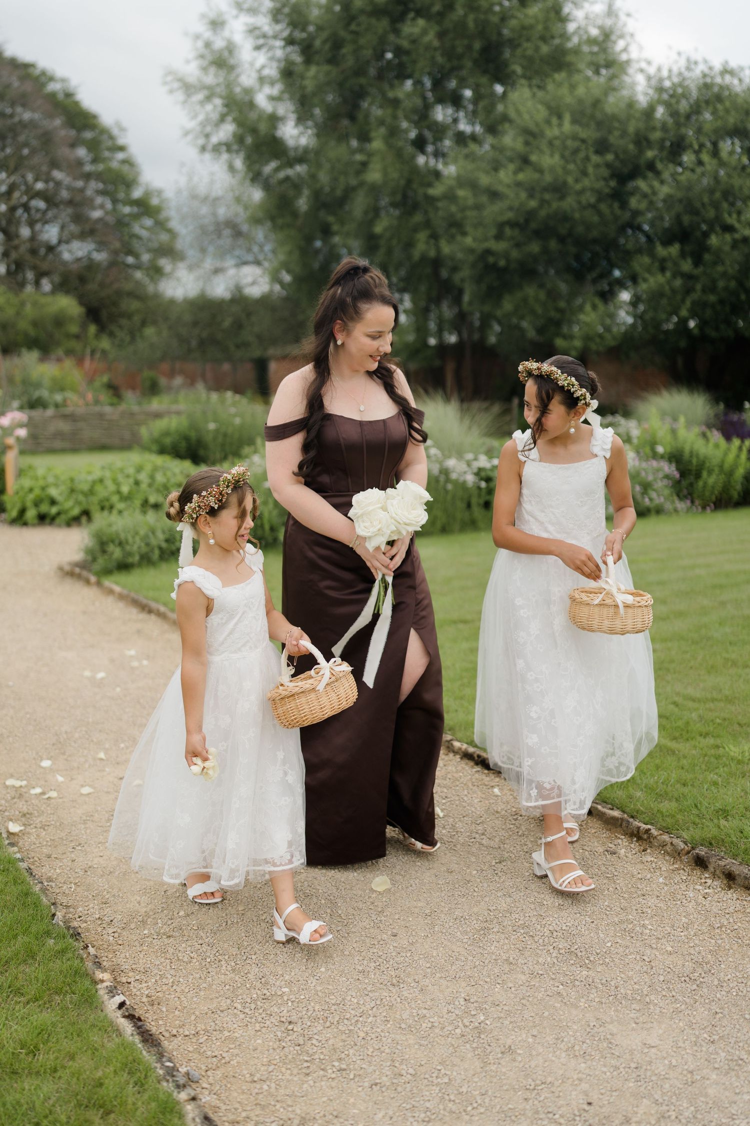 A bridesmaid in a brown gown walks with two flower girls in white dresses carrying wicker baskets.